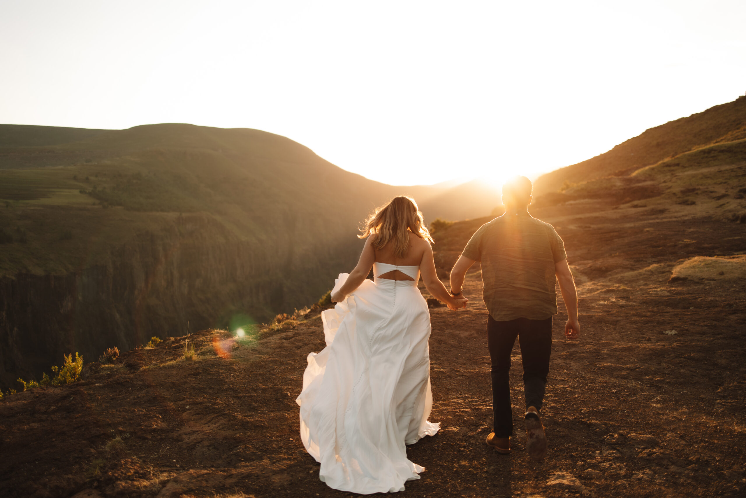 A couple holding hands walks toward the sunset on a rocky landscape, with the woman wearing a flowing white dress at Drakensburg a South Africa elopement spot