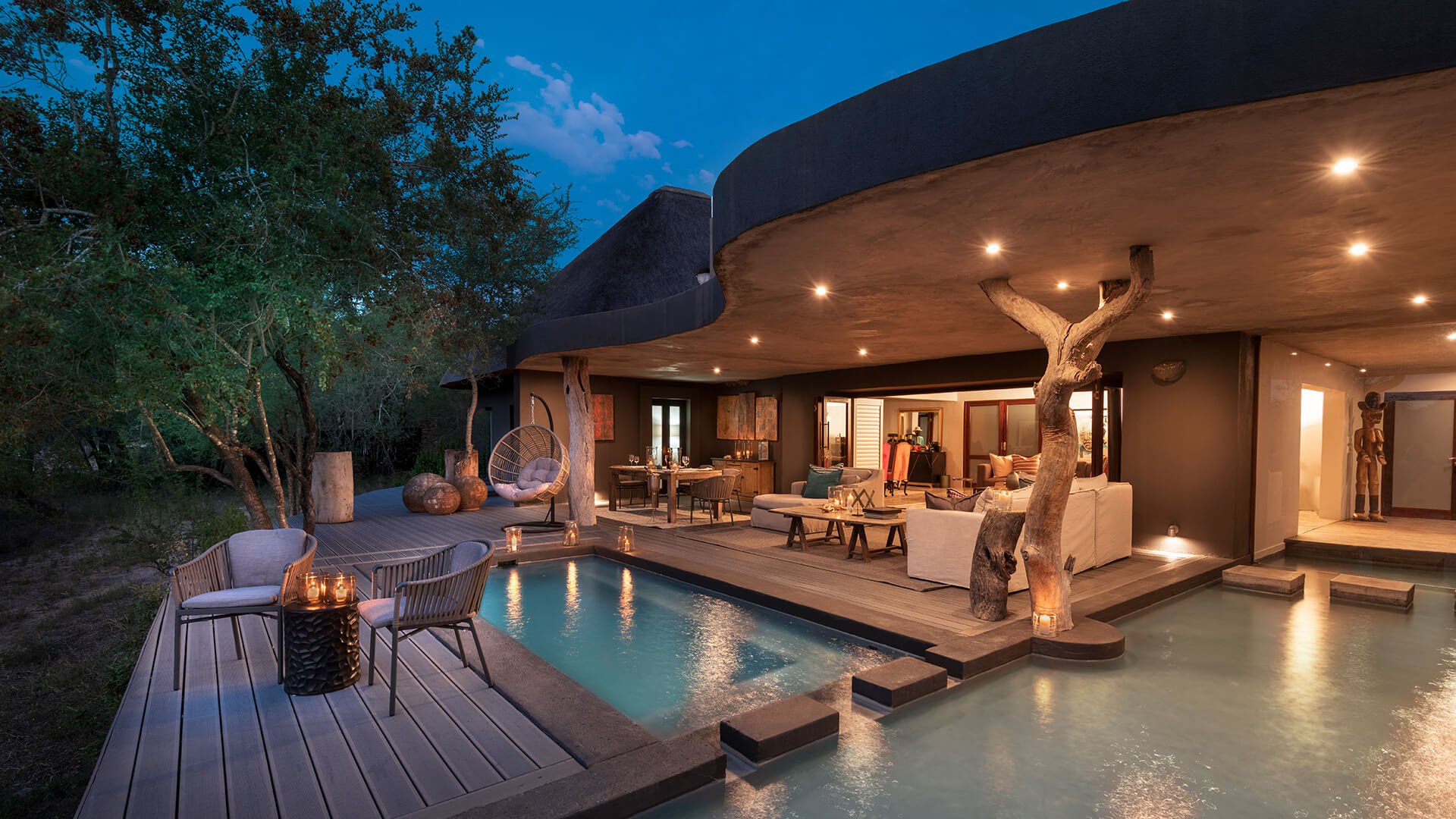 An outdoor patio with a pool at a modern house in the evening, featuring seating areas, a tree integrated into the structure, and warm lighting at Chitwa Chitwa a lodge perfect for a safari elopement