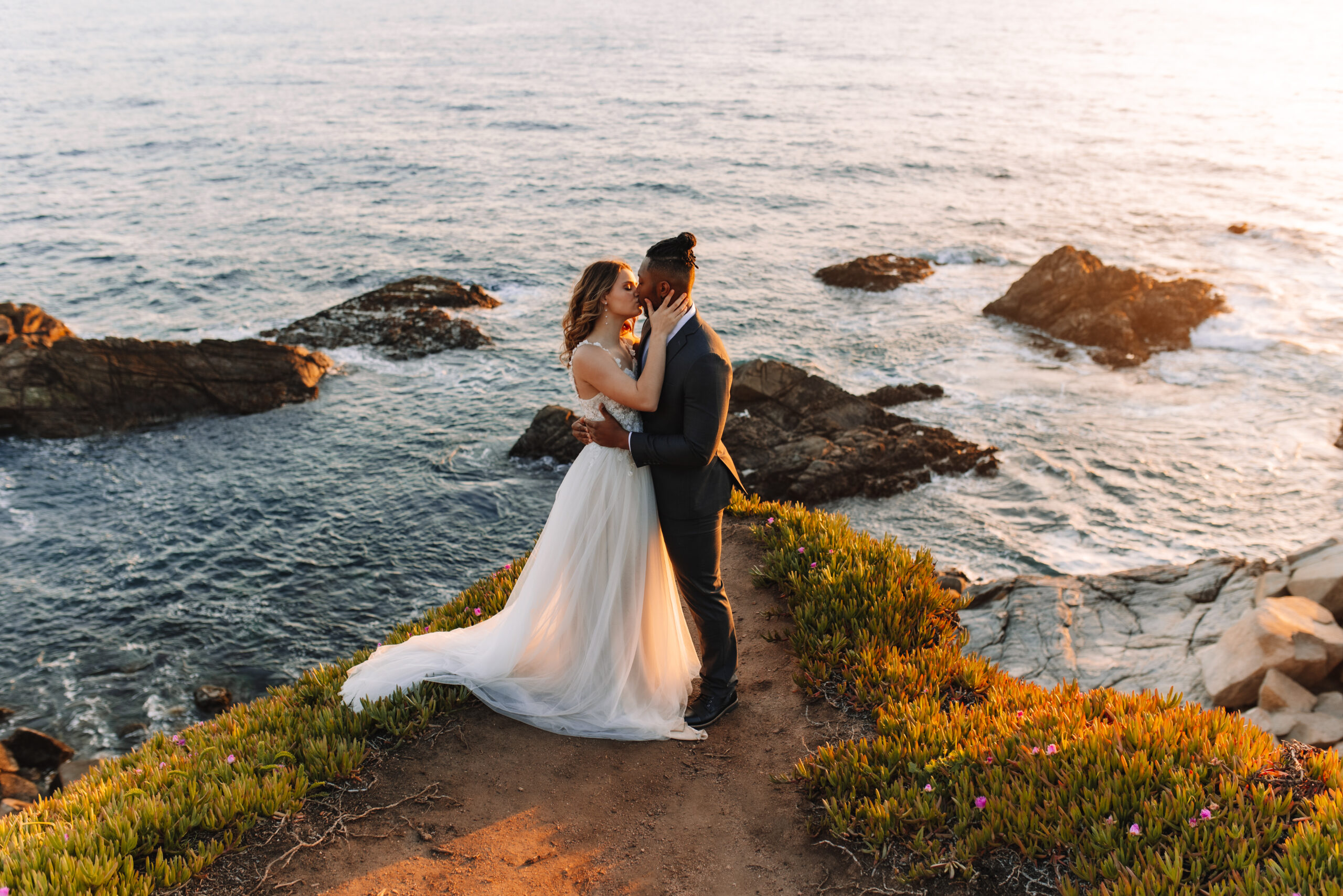 A bride and groom stand on a grassy, rocky coastal cliff at sunset, gazing into each other's eyes. The ocean and rocks are visible in the background at Chapman's Peak a South Africa Elopement Spot 