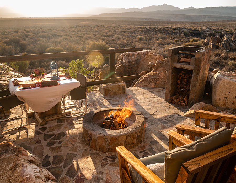Outdoor seating area with a fire pit, wooden chairs, and a table set for dining on a stone patio overlooking a vast desert landscape at sunset at Kagga Kamma Nature Reserve