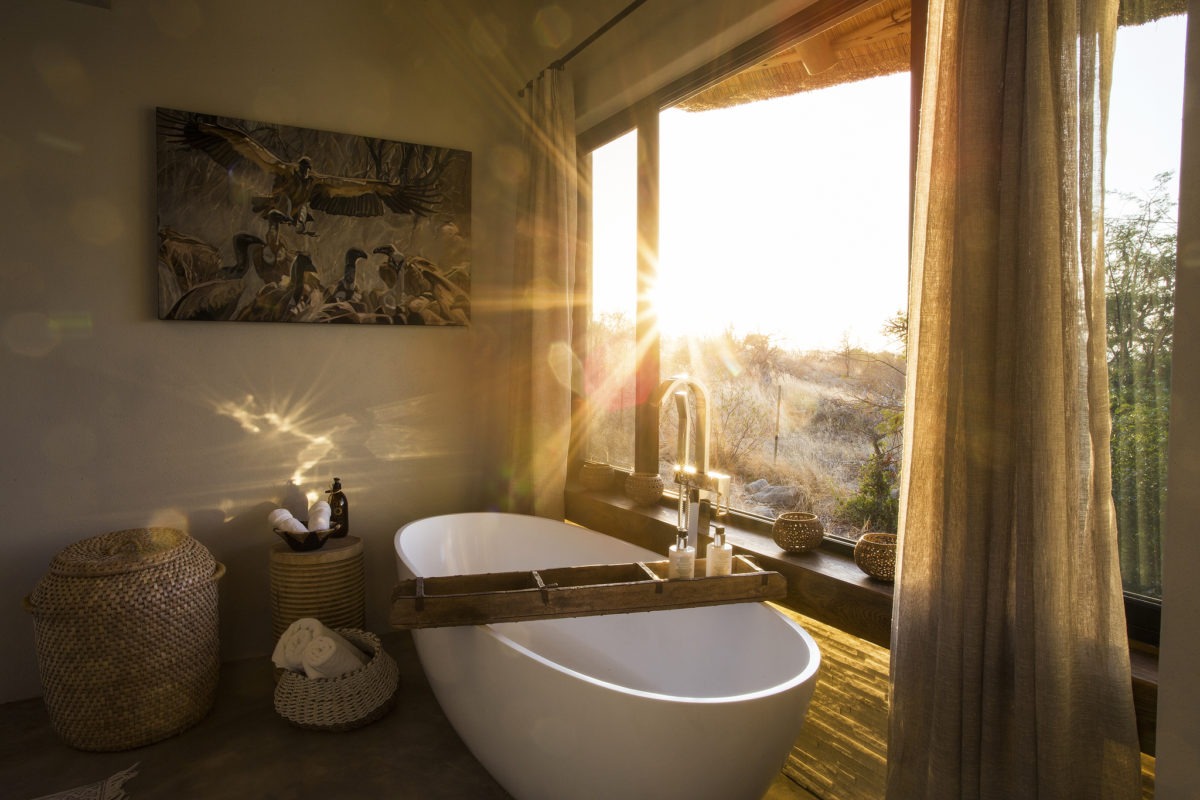 Sunlight streams into a bathroom with a freestanding bathtub, wicker basket, and large window with curtains, overlooking a natural outdoor setting at Misava Safari Camp a perfect lodge for a safari elopement!