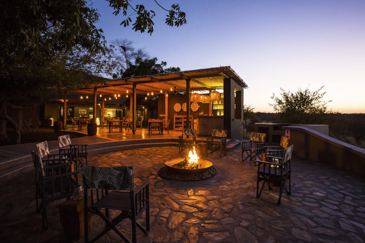 Outdoor seating area with a lit fire pit, surrounded by wooden chairs, and an open-air building with string lights at dusk. Trees and shrubs are visible in the background at Misava Safari Camp a perfect lodge for a safari elopement!