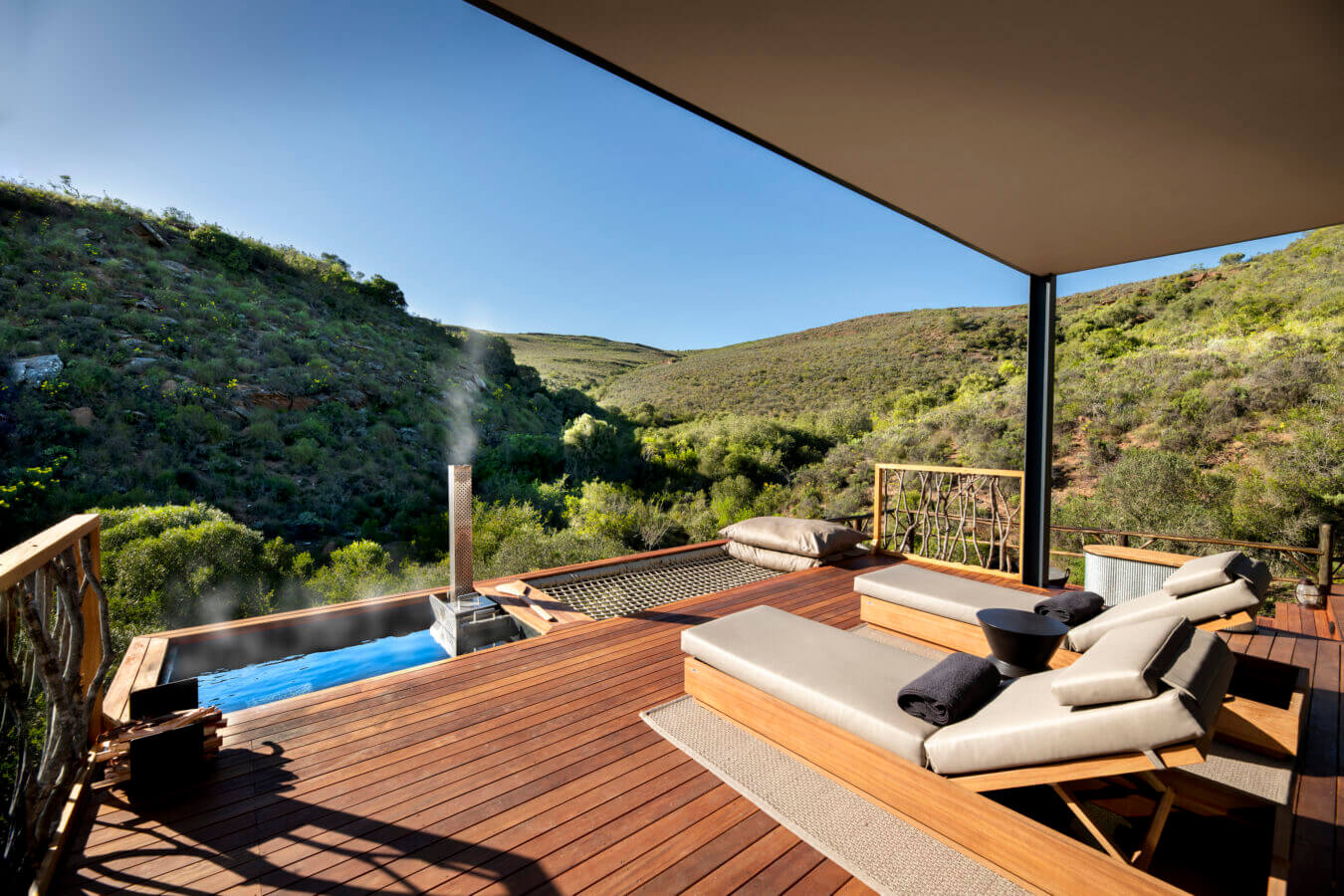 A wooden deck with lounge chairs, a small pool emitting steam, and a hammock, overlooks a green hilly landscape under a clear blue sky at Melozhori Private Game Reserve in south africa