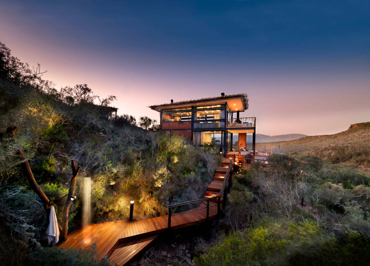 A modern home with large glass windows, surrounded by greenery, under a twilight sky. A wooden walkway leads to the entrance, illuminated by small lights at Melozhori Private Game Reserve