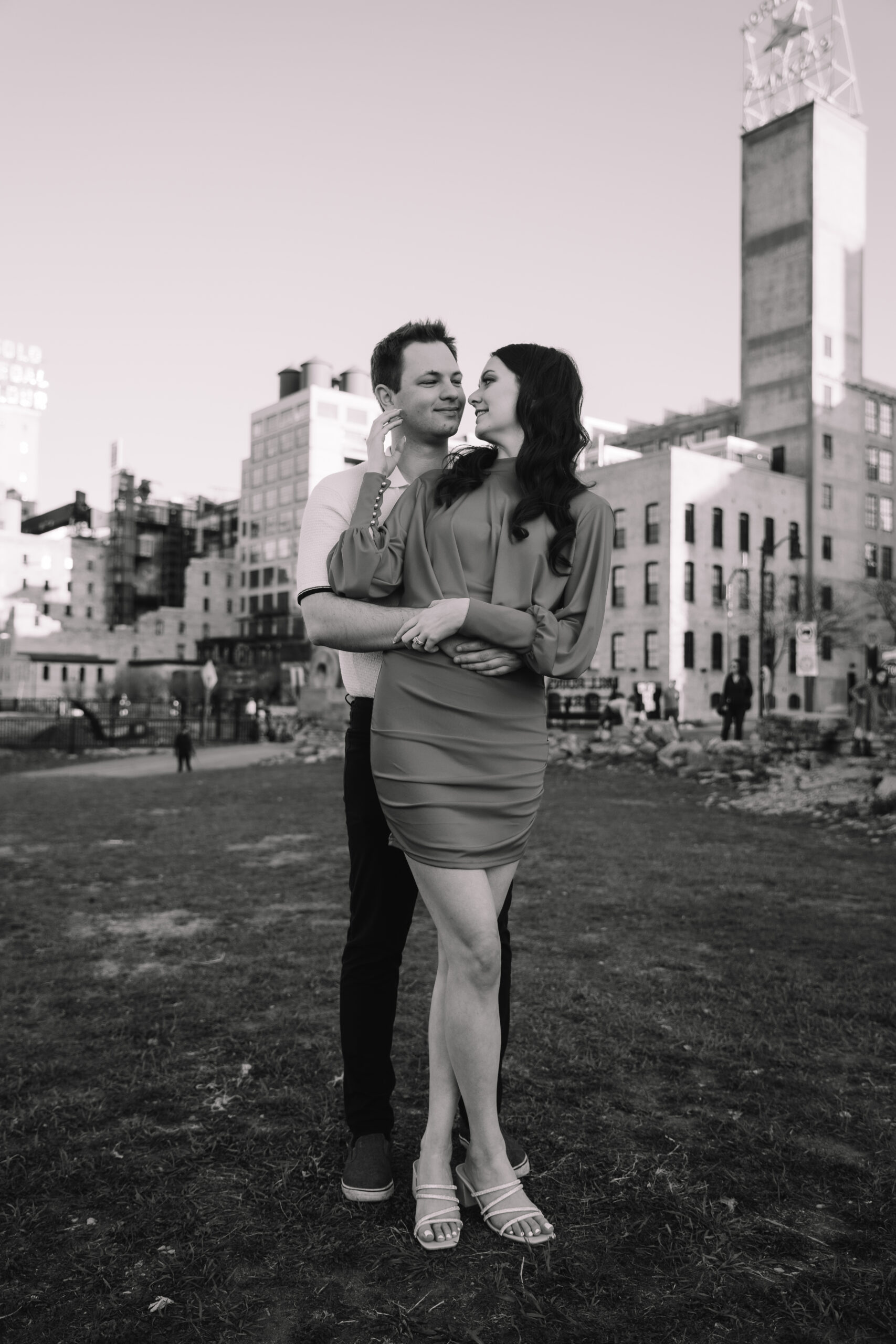 A man and woman stand together in an urban park with tall buildings in the background. The man stands behind the woman, both smiling, as he embraces her in a pose. The scene is in black and white.