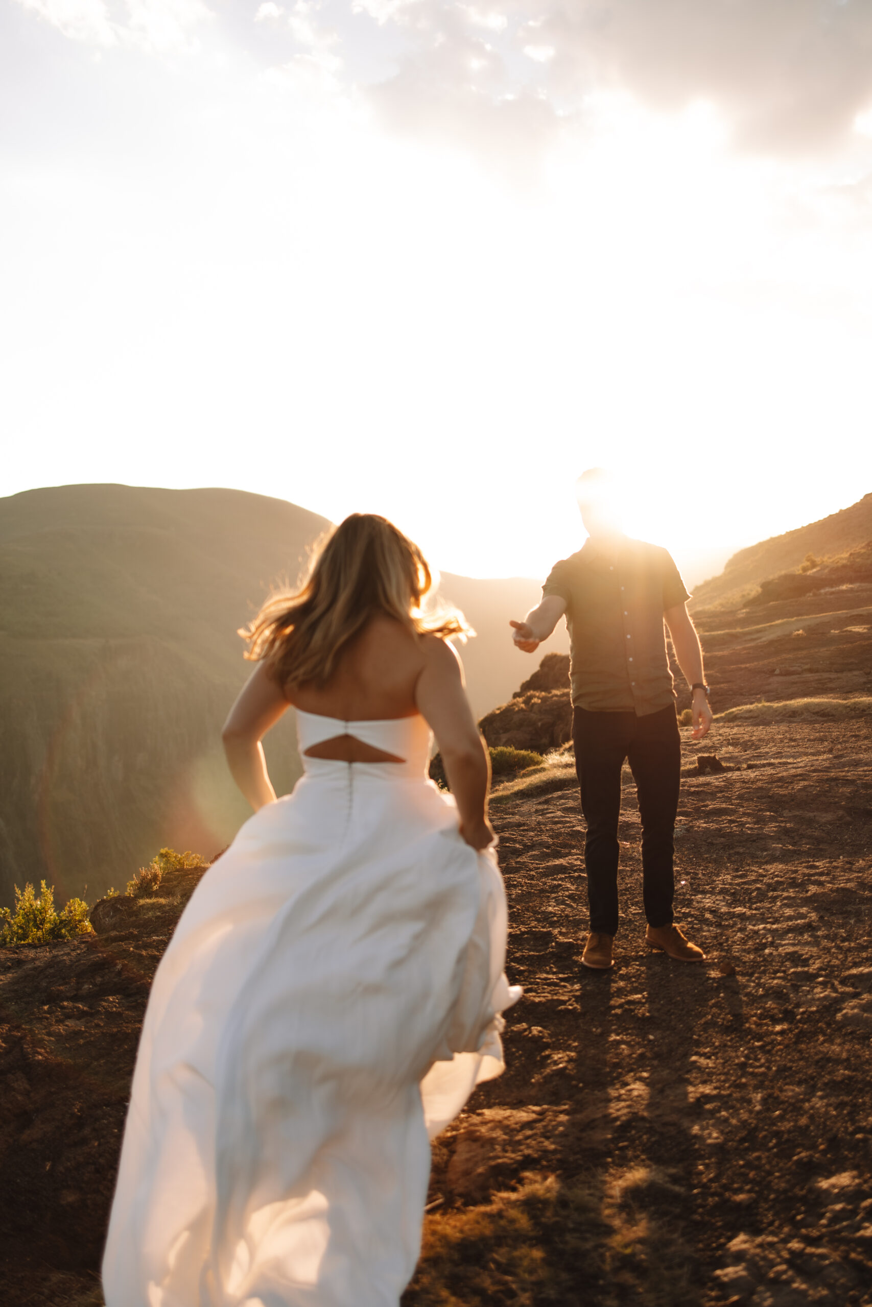 A woman in a white dress runs toward a person in casual attire at sunset, on a rocky hilltop with mountains in the background at a south africa elopement spot