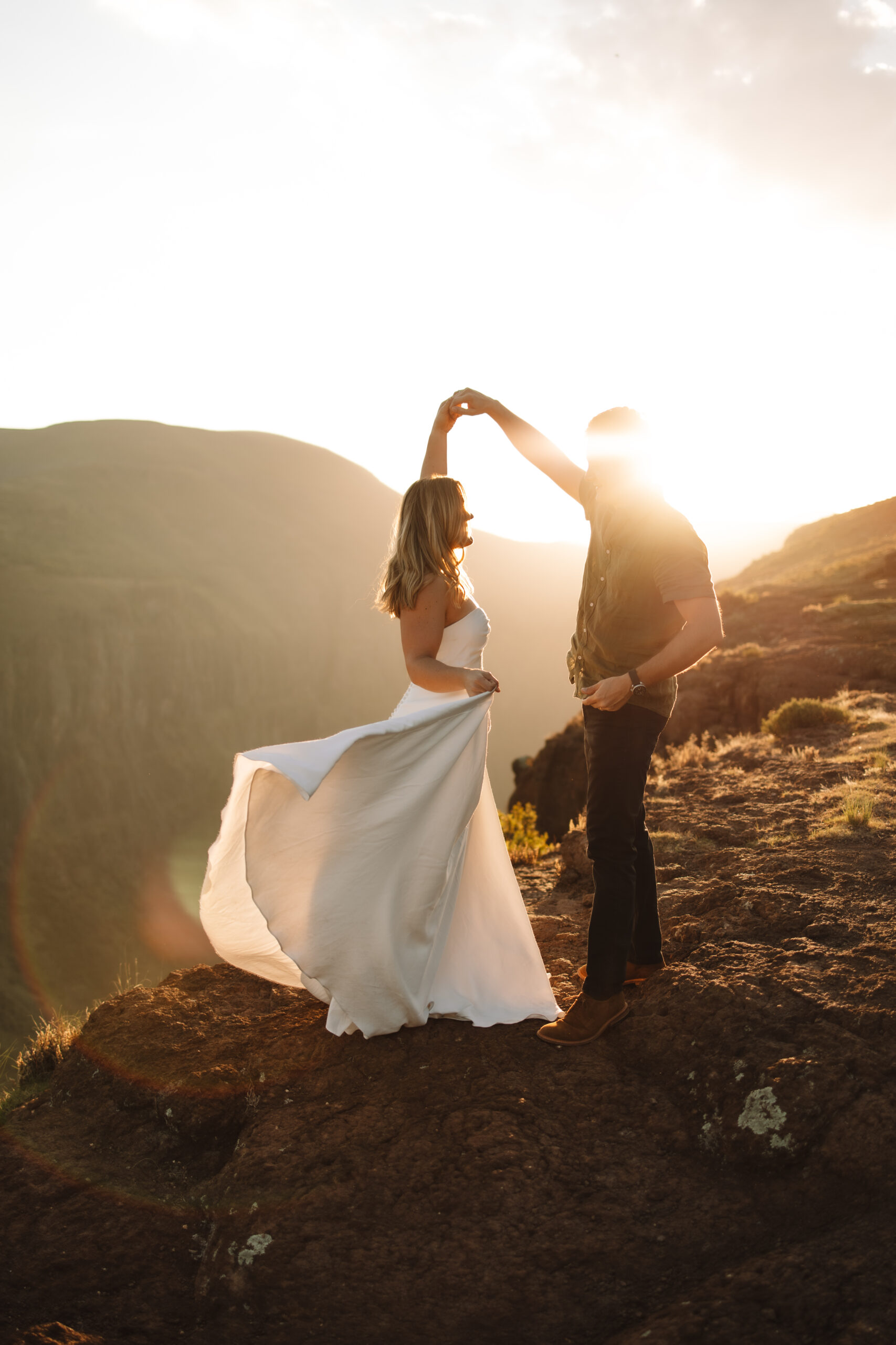 A woman in a white dress runs toward a person in casual attire at sunset, on a rocky hilltop with mountains in the background at a south africa elopement spot