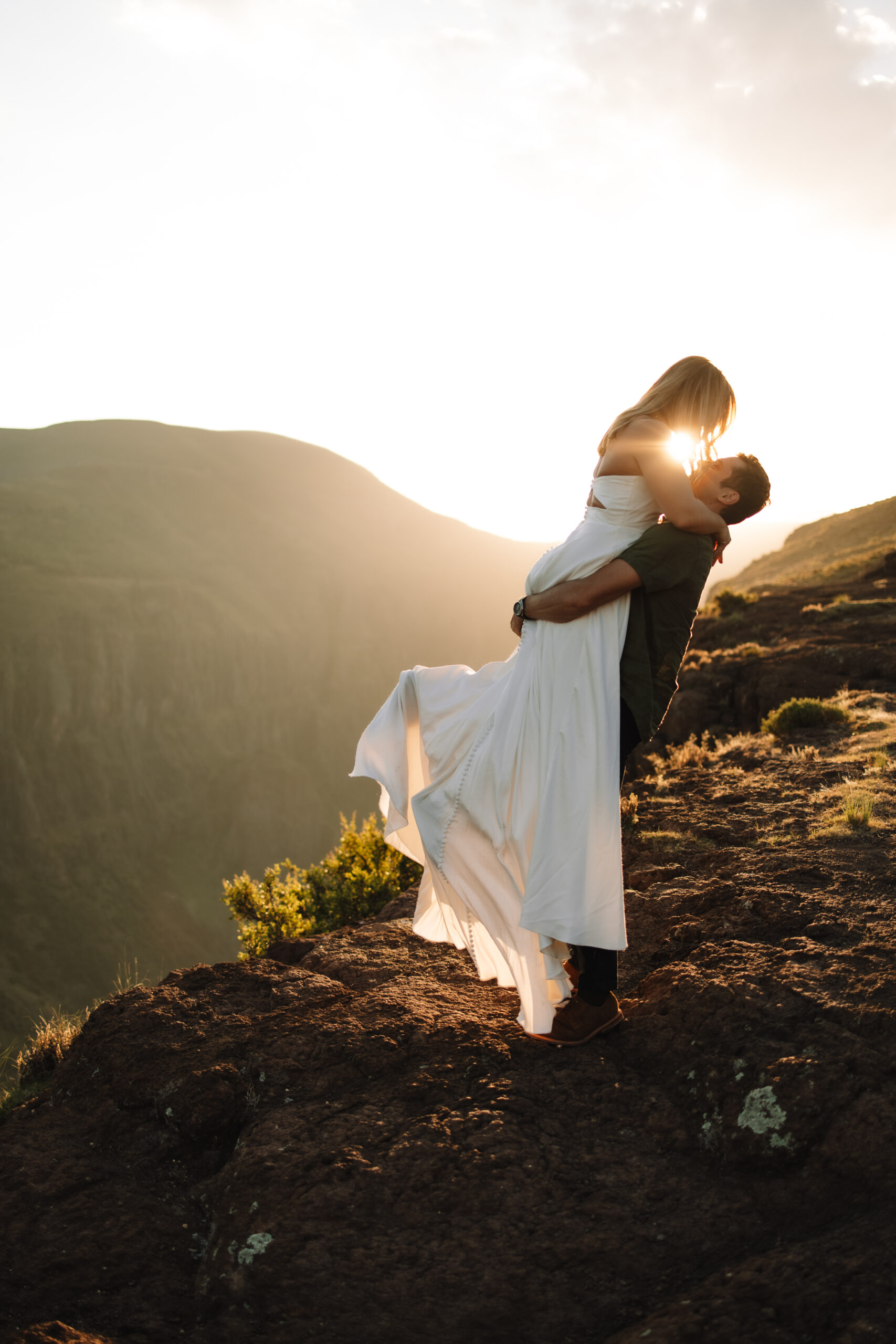 A woman in a white dress runs toward a person in casual attire at sunset, on a rocky hilltop with mountains in the background at a south africa elopement spot