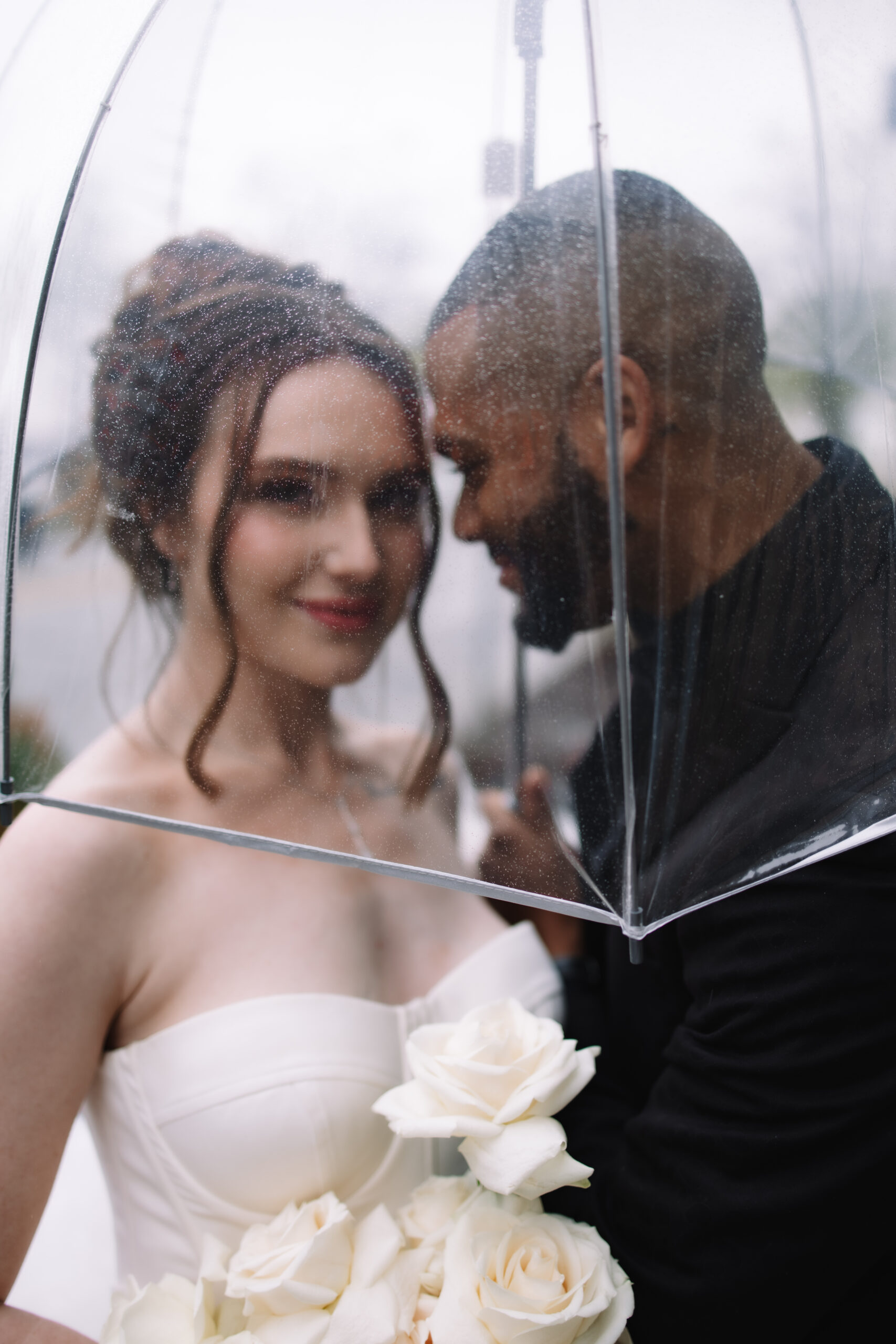 A bride and groom stand under a clear umbrella on a rainy day, sharing a close moment. The bride holds white roses and wears a strapless dress.