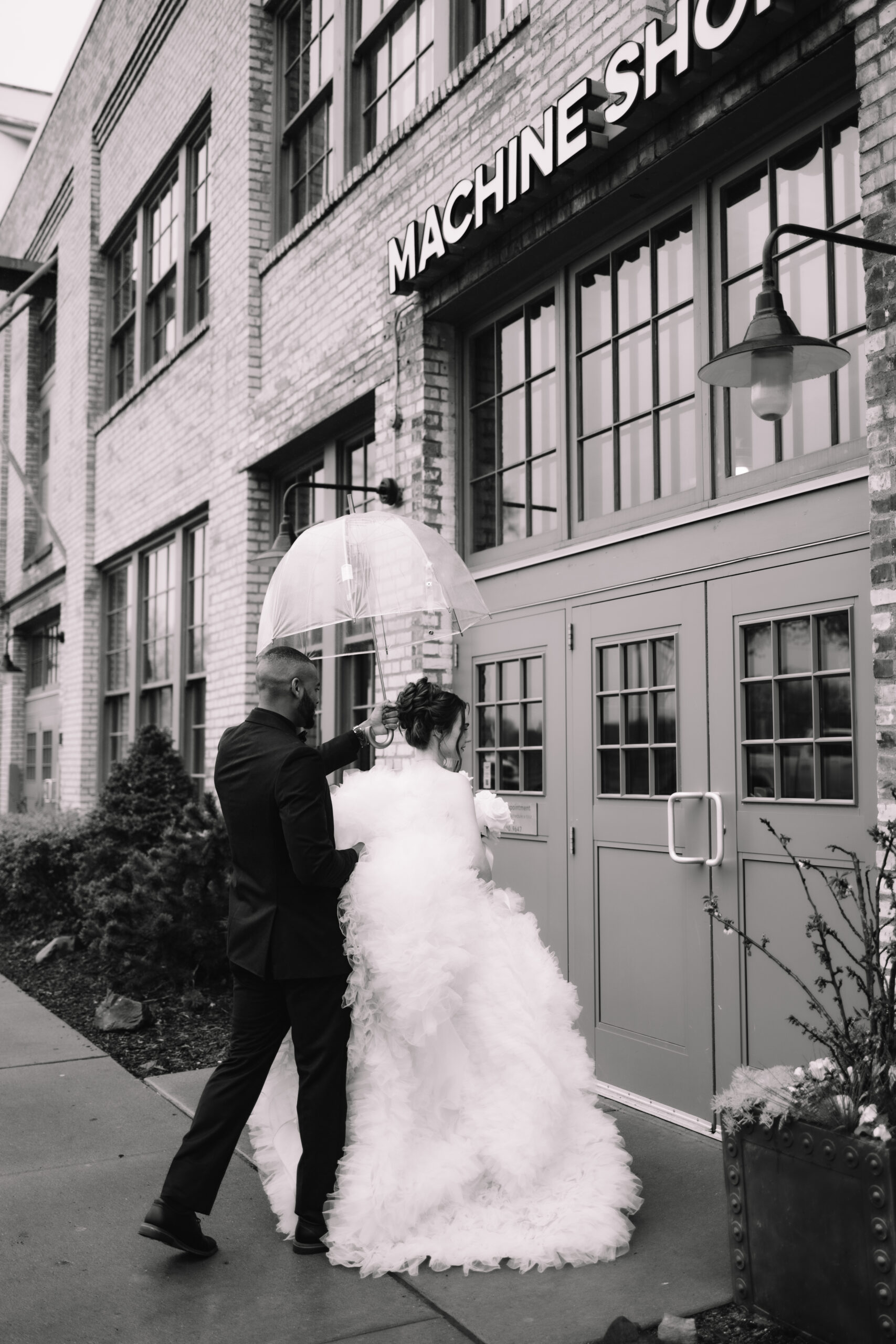 A couple under an umbrella stands by the door of a brick building labeled "Machine Shop." The bride is in a voluminous dress, and the groom wears a suit at The Machine Shop Minneapolis