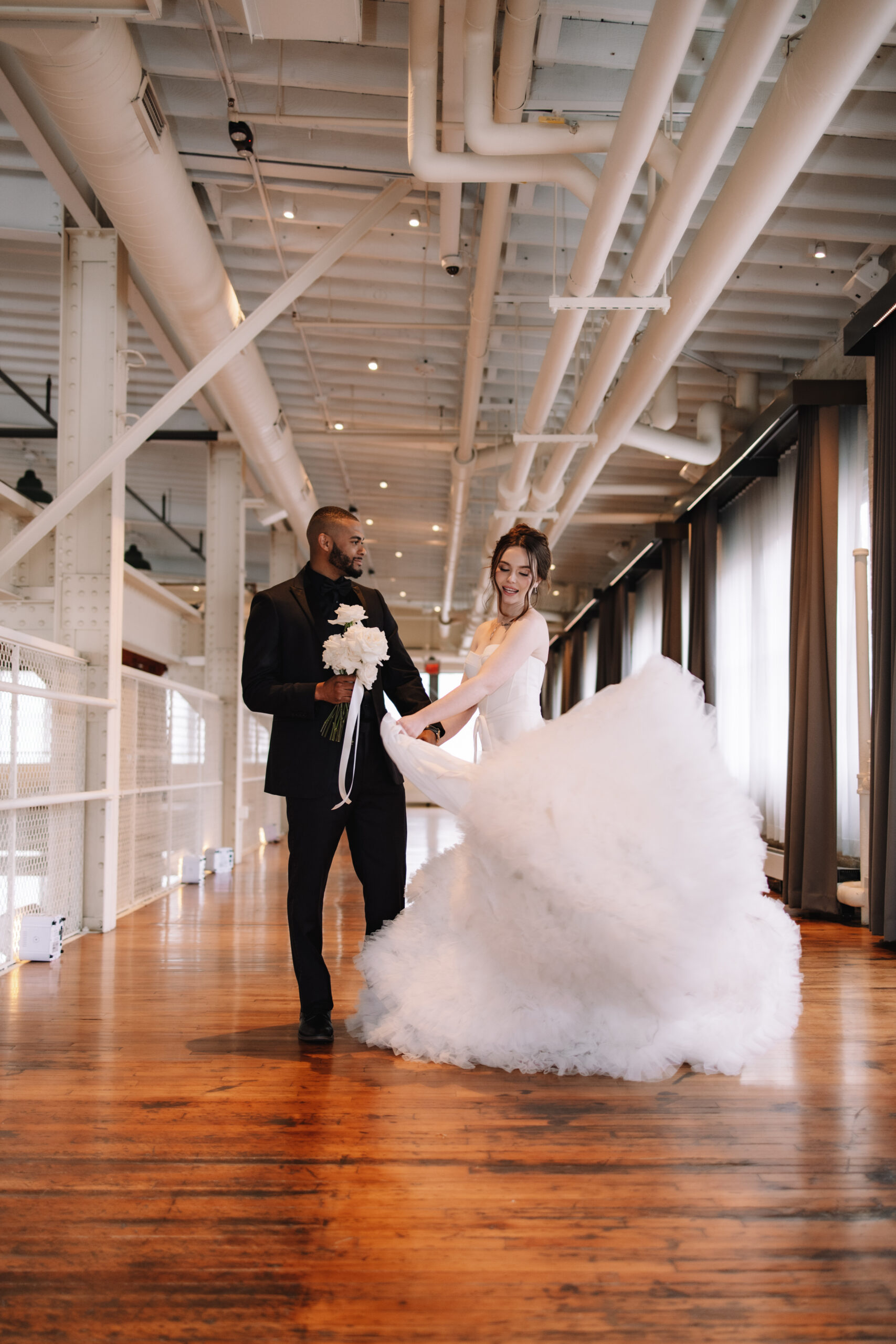 A couple in wedding attire dances in a bright, industrial-style venue. The bride's gown flares out as she twirls, and the groom holds a bouquet of flowers in The Machine Shop in Minneapolis
