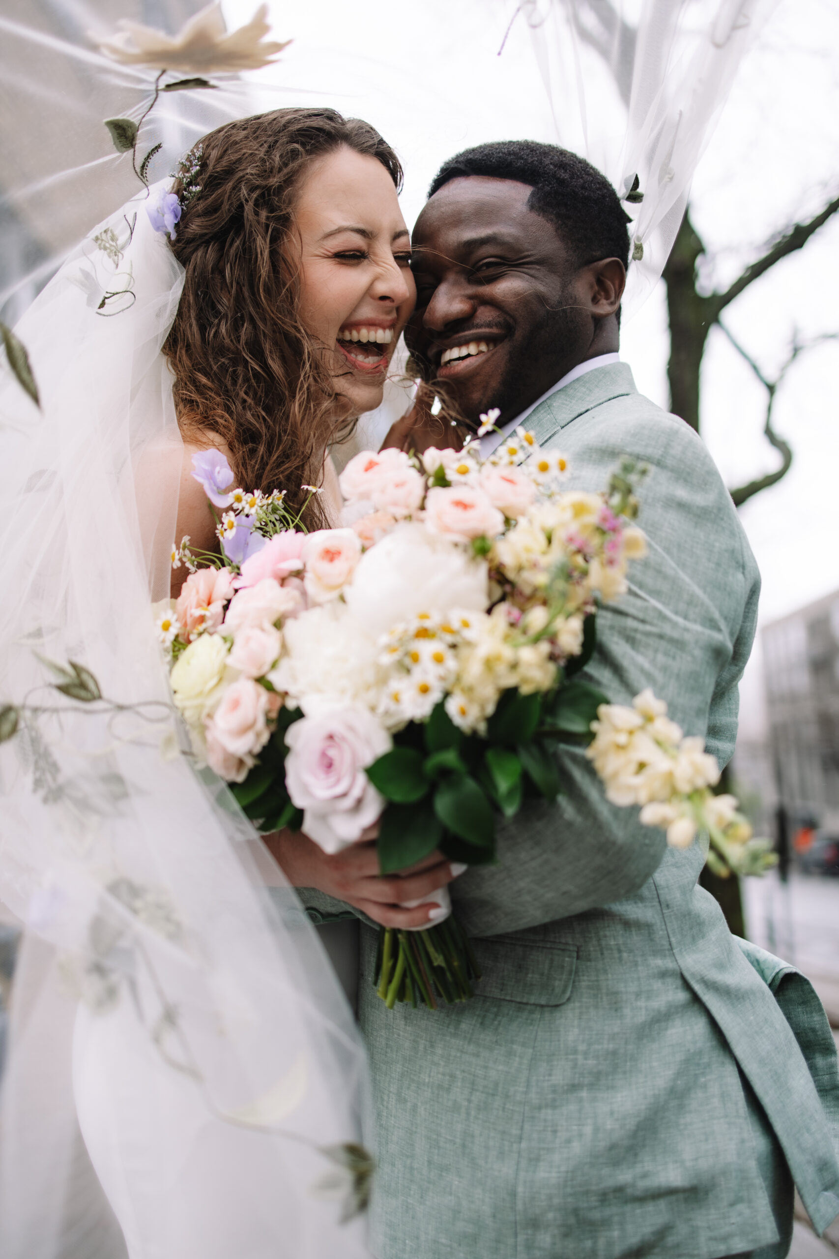 A bride and groom smile joyfully while embracing each other and holding a colorful bouquet of flowers.
