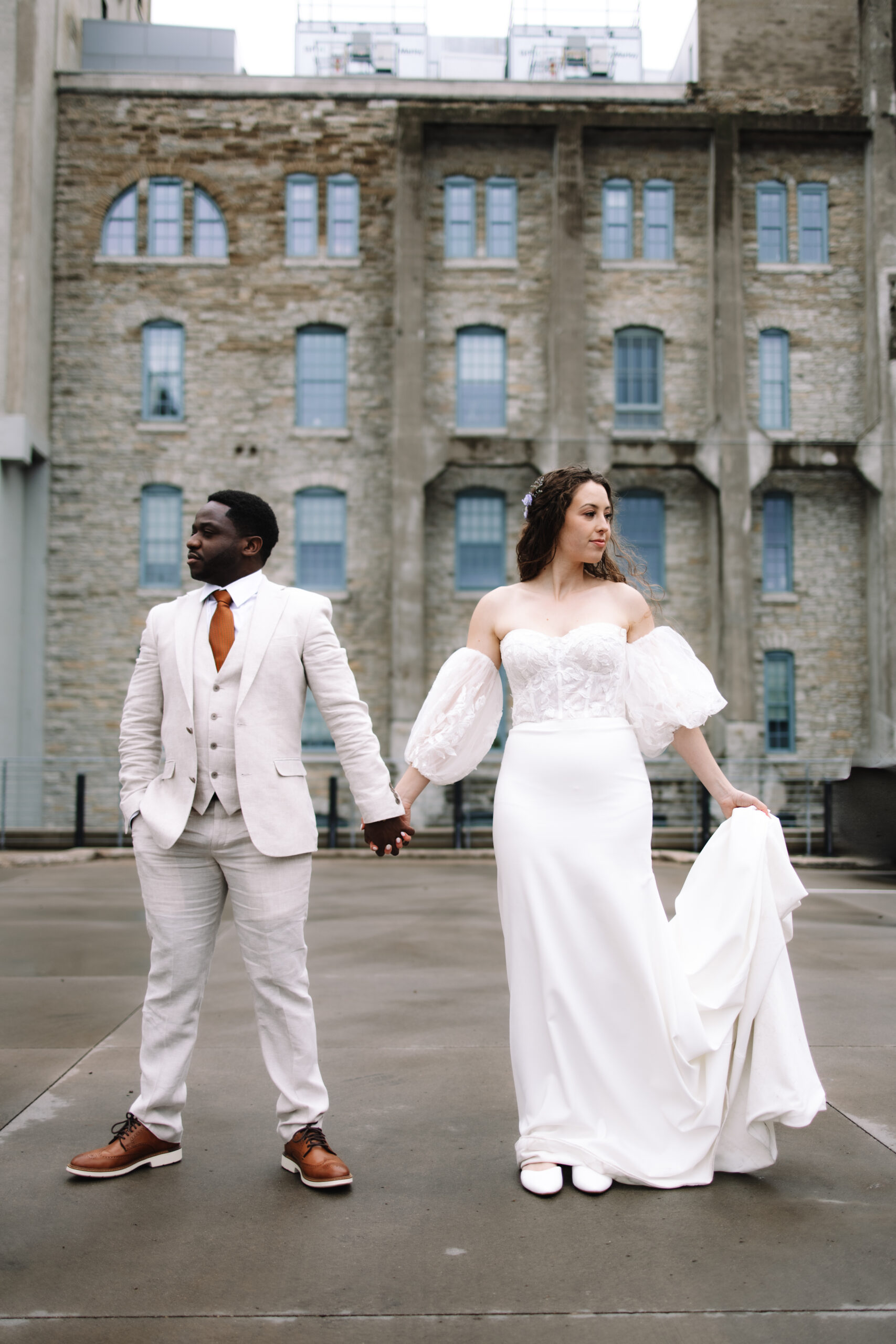 A bride and groom standing next to each other holding hands, looking in opposite directions with a large brick building behind them at the machine shop in Minneapolis
