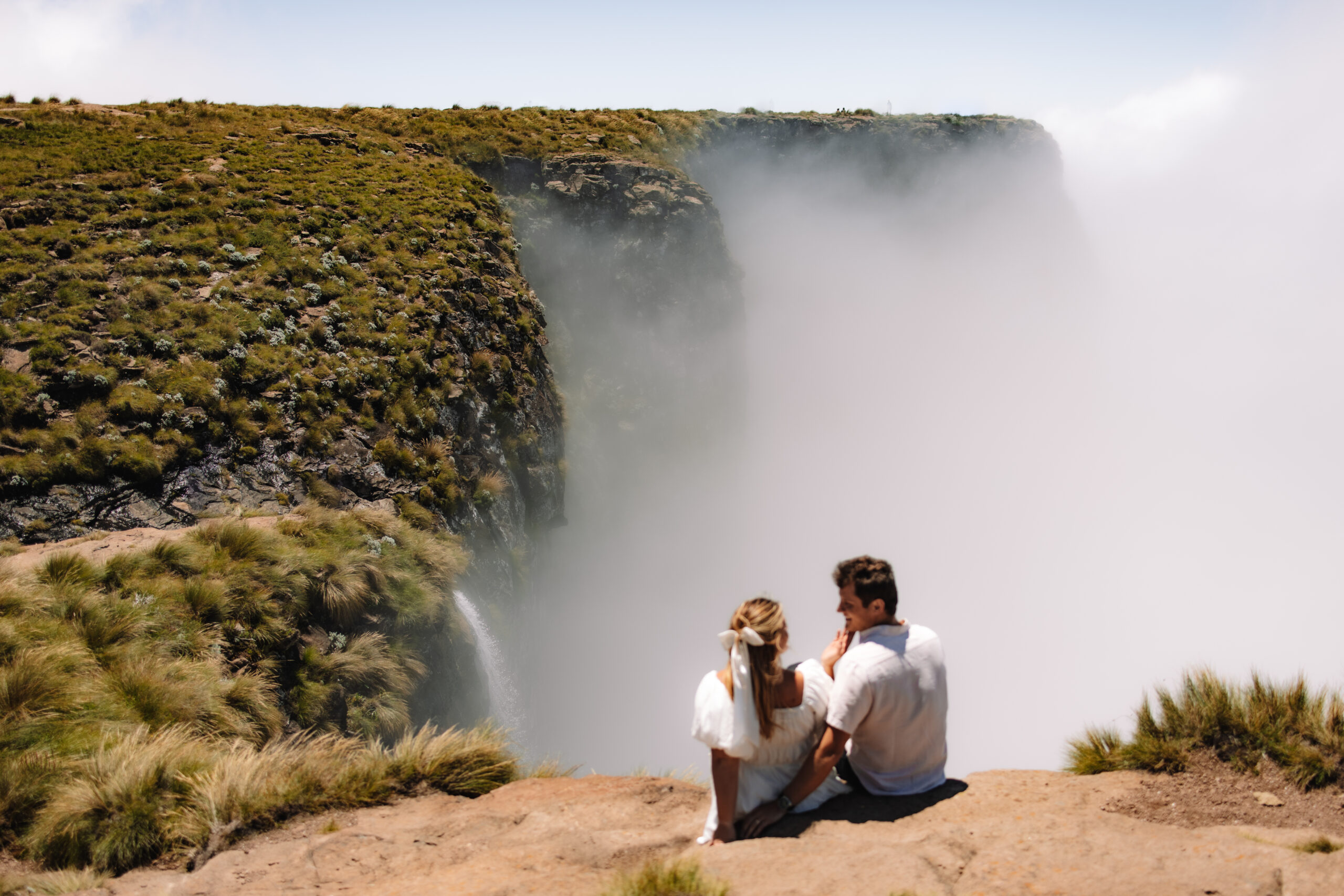 Two people sit close together on a rocky edge, overlooking a misty waterfall surrounded by grass and foliage at Tugela Falls a South Africa Elopement spot 