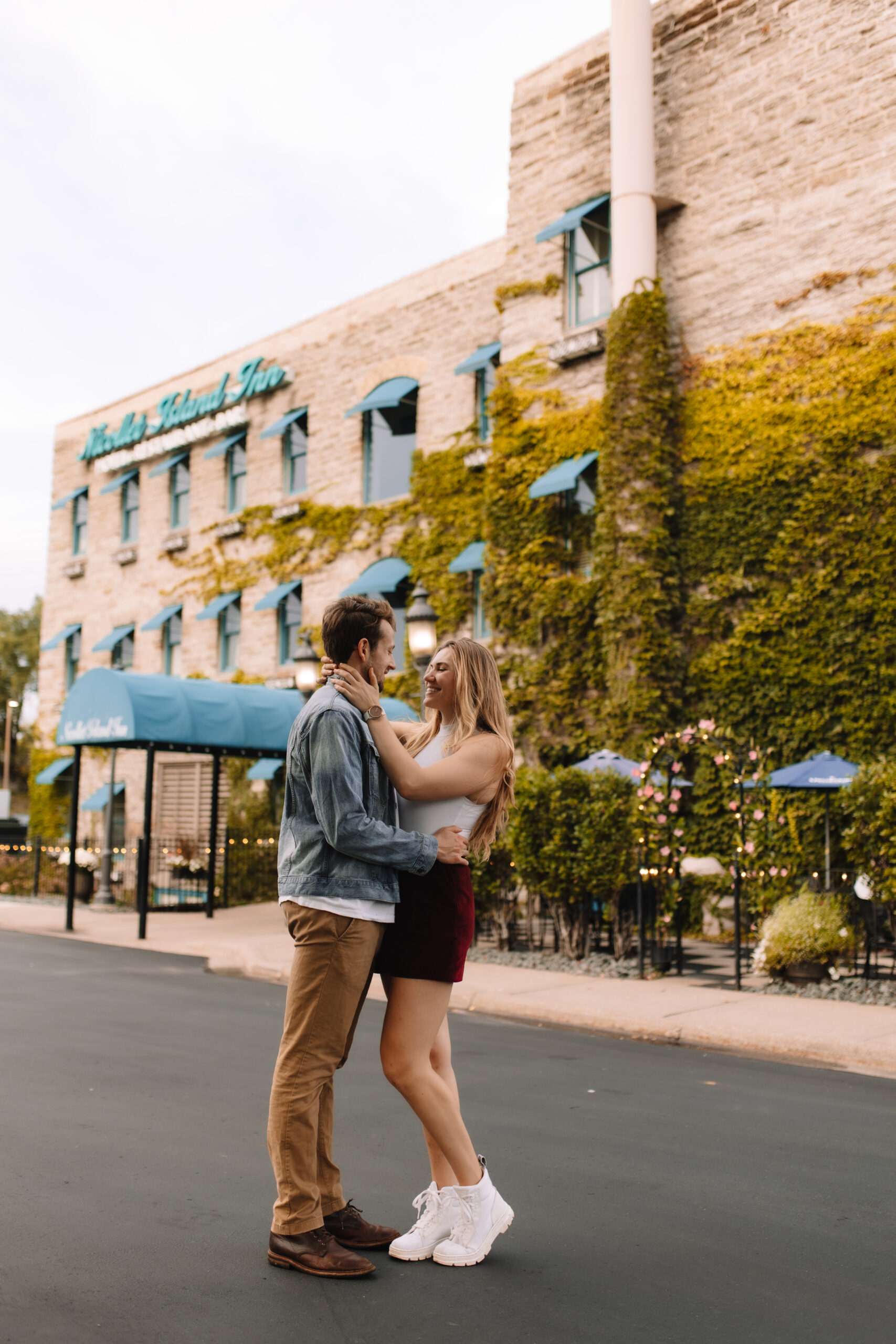 A couple stands close together, gazing at each other affectionately, in front of a stone building with ivy and blue awnings.