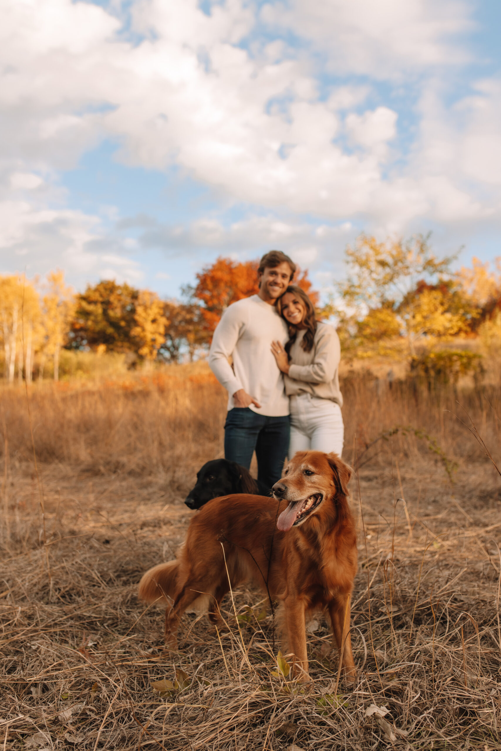 Newly engaged couple smiling in a field of grass with their pup in fall engagement outfits