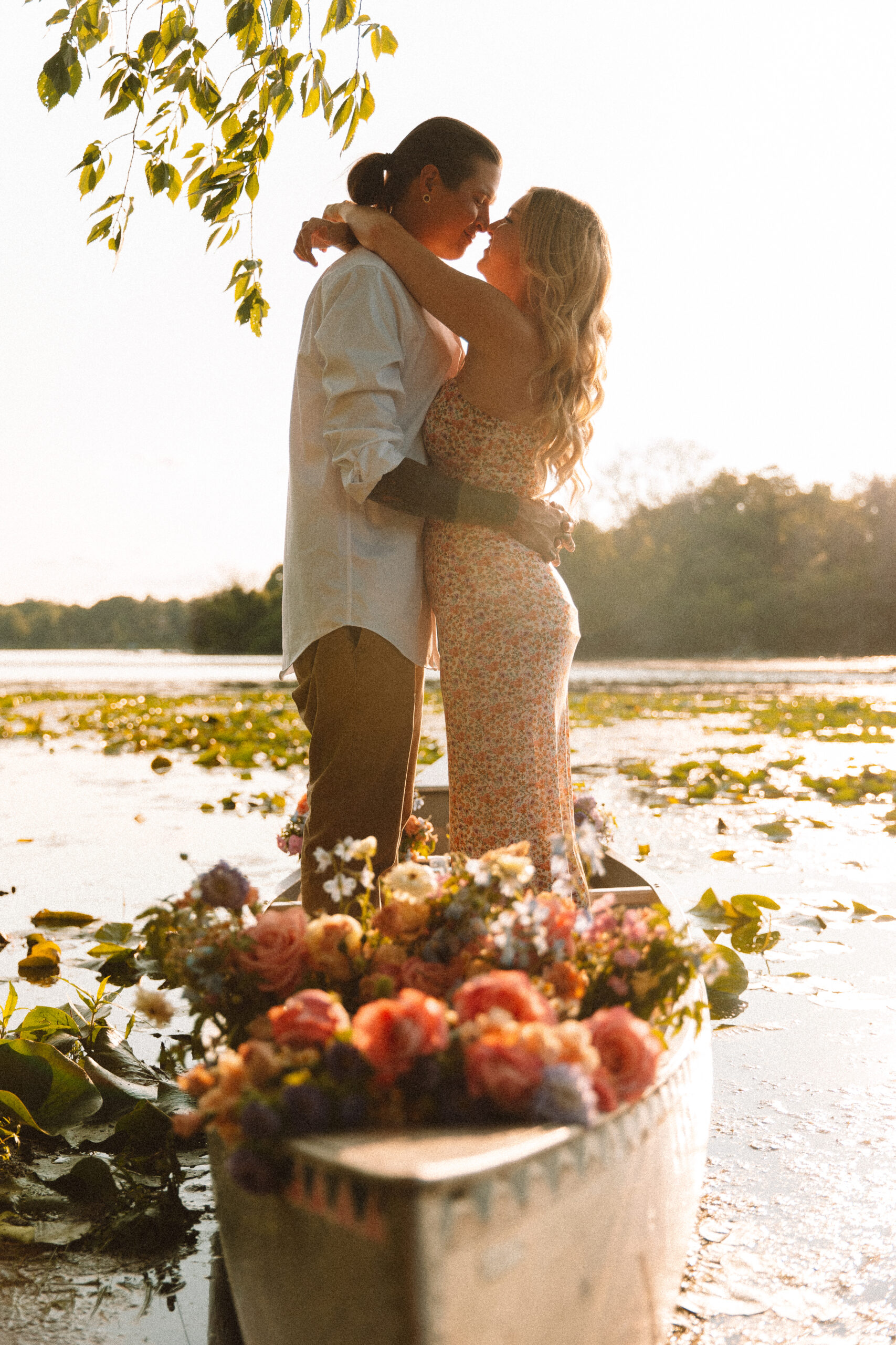 Newly engaged couple standing in a canoe full of flowers, wearing a long maxi dress and white button up and pants