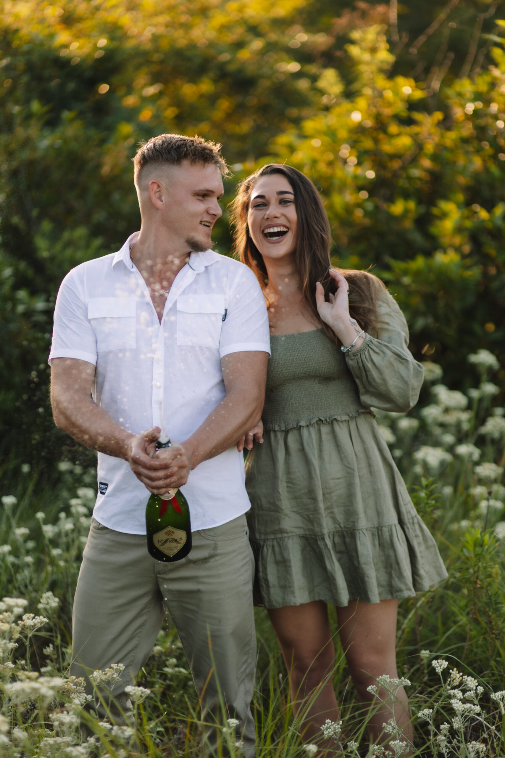 Engaged couple wearing cute engagements in a flower field popping champagne, perfect inspiration to help you on what to wear for your engagement photos