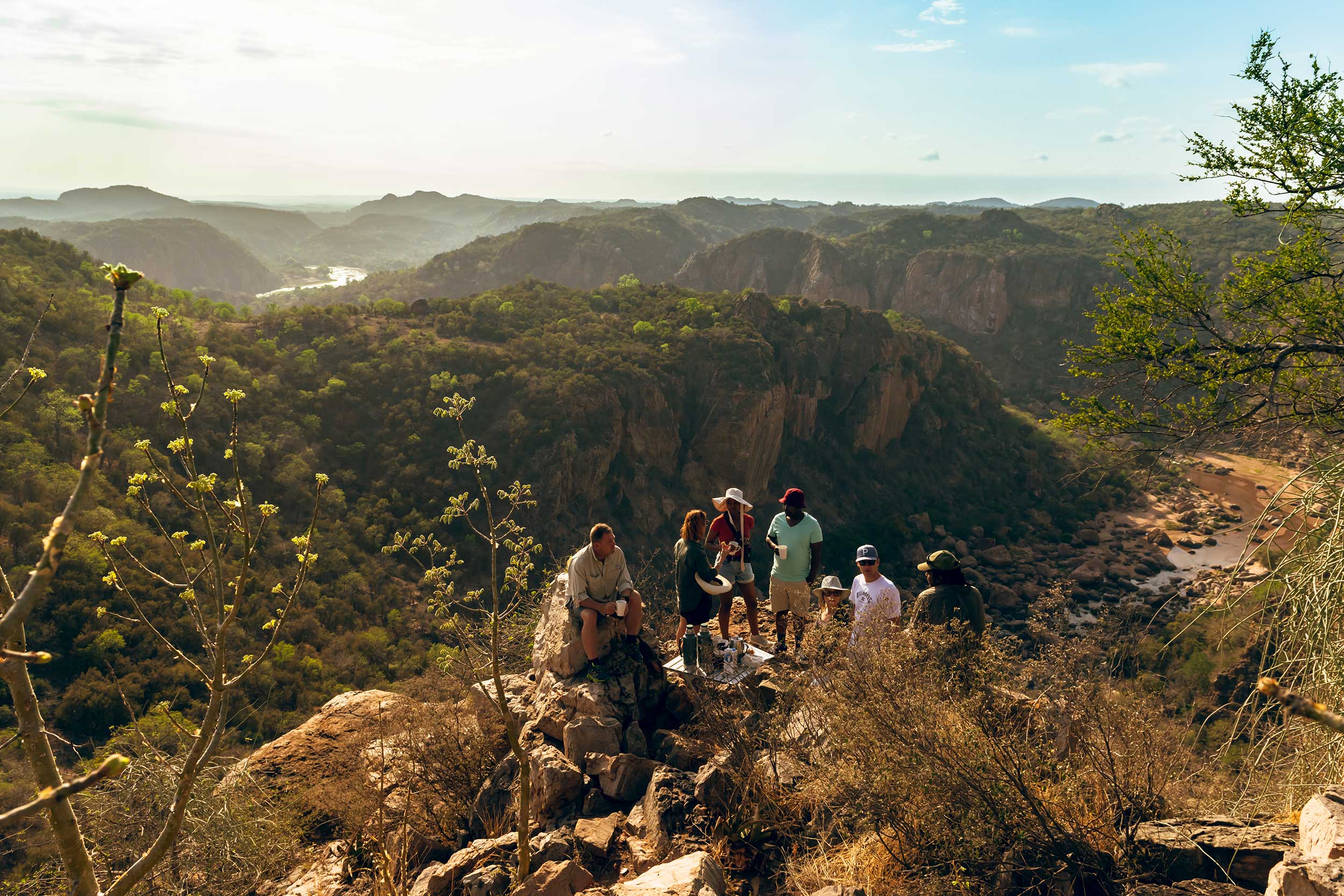 Group of people hiking on a rocky ridge with a scenic view of a lush valley, mountains, and a winding river in the background under a sunny sky