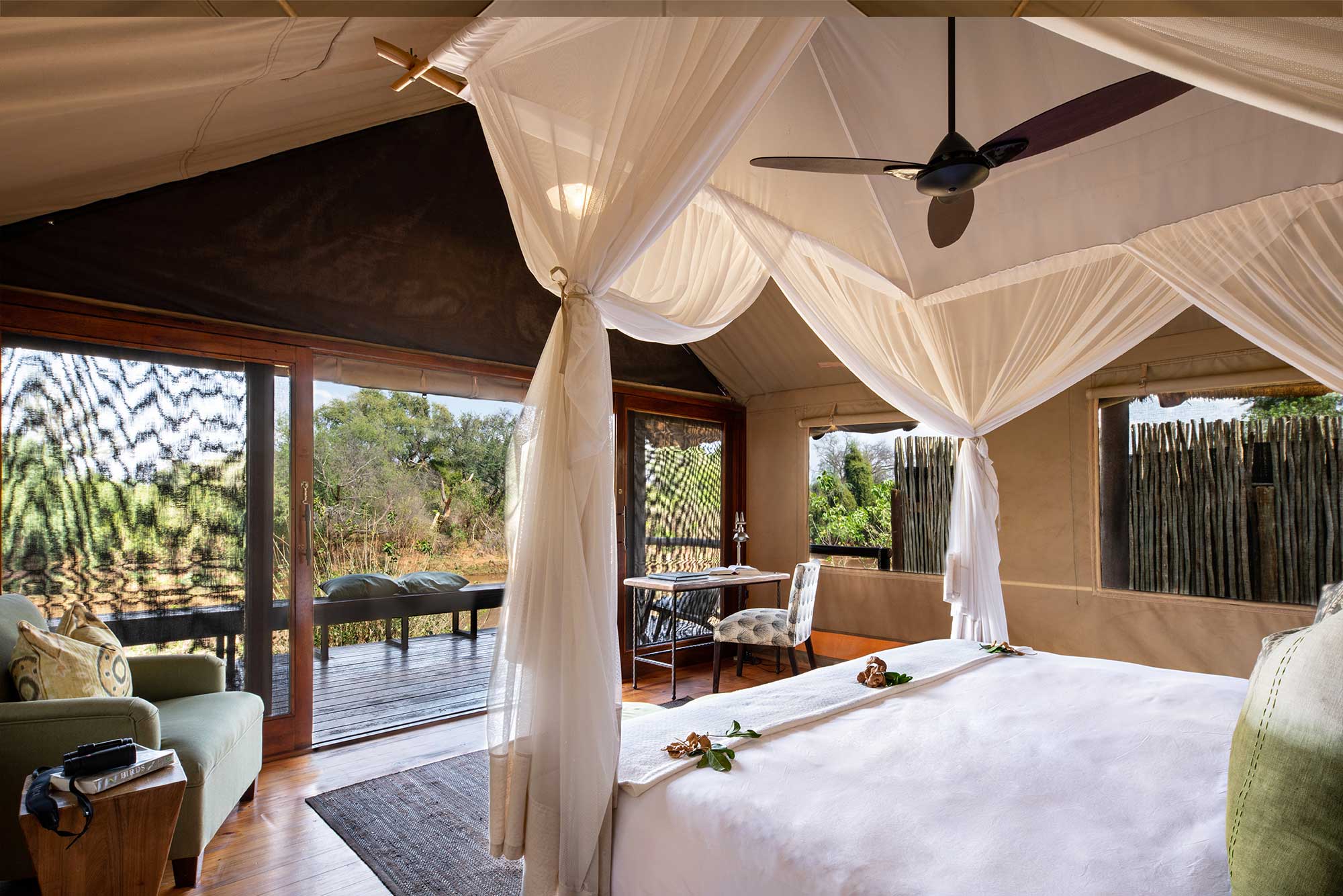 A luxury tent interior with a canopy bed, white linens, a ceiling fan, a writing desk, a green sofa, and an open view of a wooden deck and lush greenery through large glass doors at return Africa a lodge for a safari elopement
