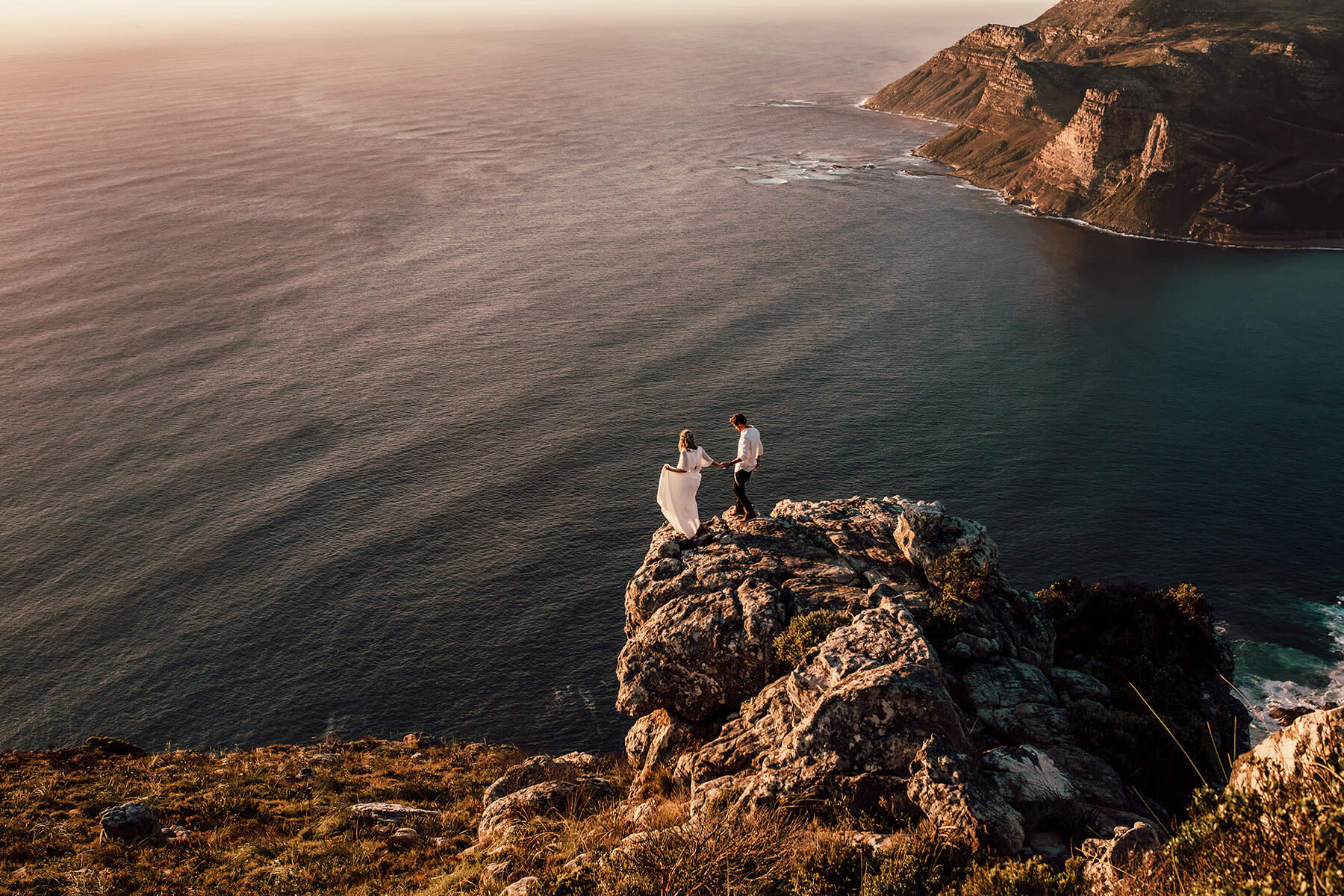 A bride and groom stand on a grassy, rocky coastal cliff at sunset, gazing into each other's eyes. The ocean and rocks are visible in the background at Chapman's Peak a South Africa Elopement Spot