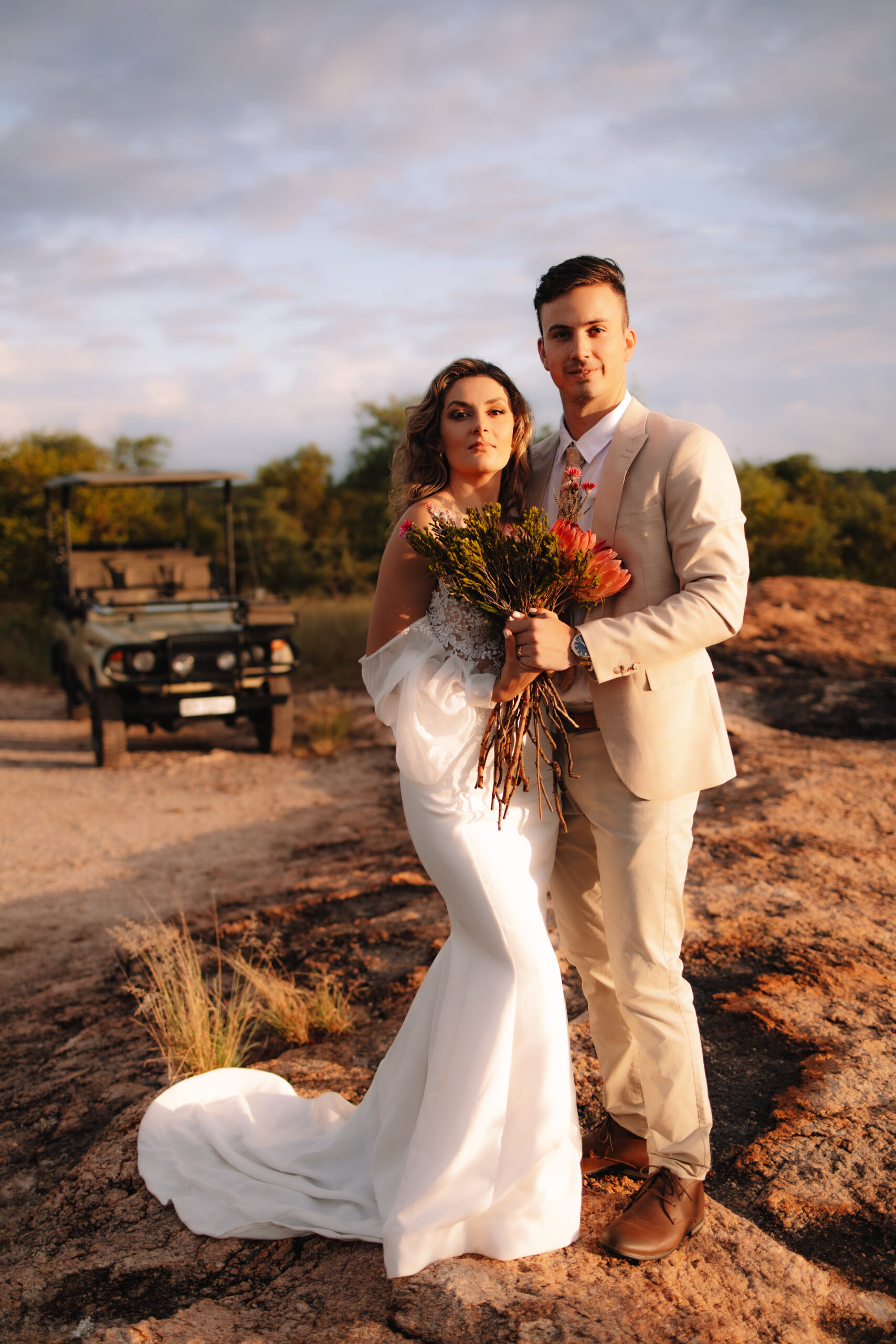 A bride and groom standing next to each other on a cliff in Kruger National Park holding a protea bouquet with a safari vehicle behind them