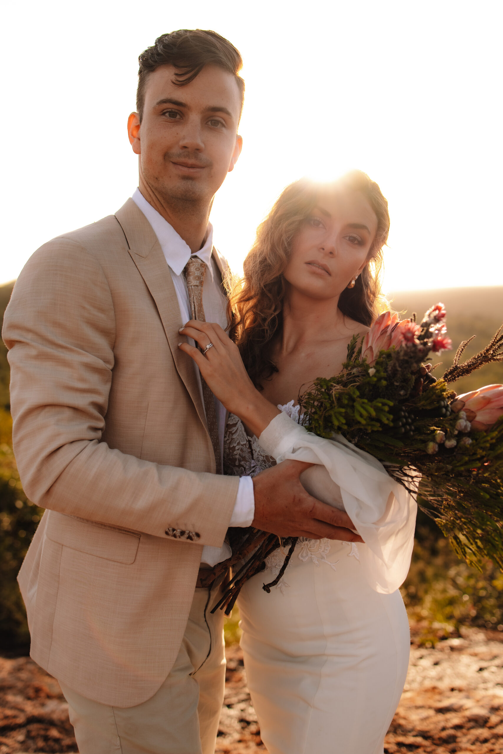 A couple embraces on a rocky ledge at sunset. The woman wears a white off-shoulder gown, and the man is in a beige suit. Forested hills extend into the distance in South Africa on their safari elopement
