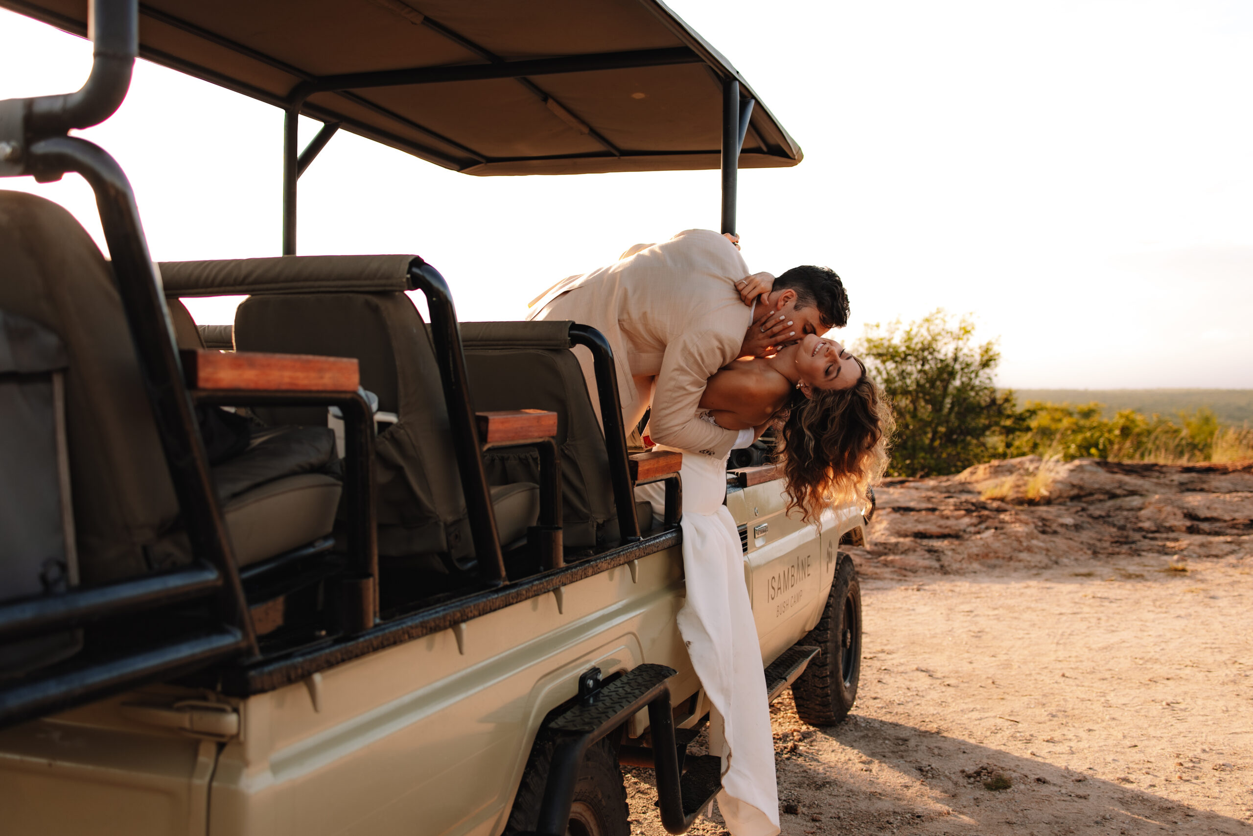 Safari elopement couple hanging out of the safari vehicle in their wedding attire while groom kisses bride's neck