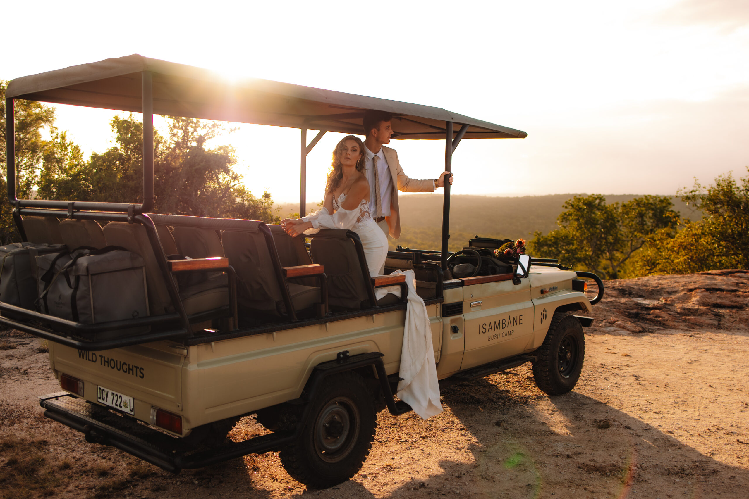 A couple dressed in wedding attire rides in an open-top safari vehicle at sunset, parked on a dirt path overlooking a scenic landscape in South Africa