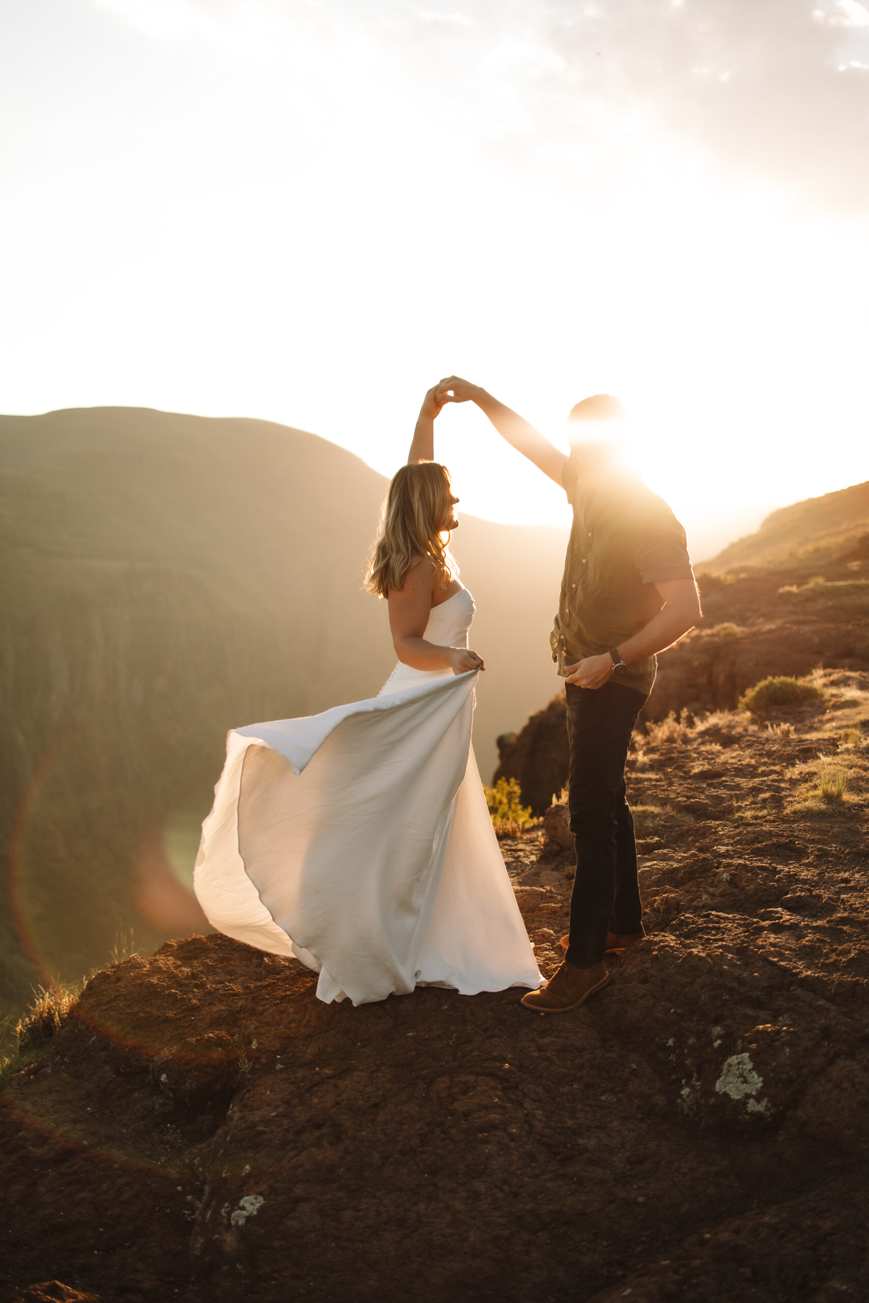 Elopement couple dancing on the edge of a cliff in South Africa as the sun sets behind the rolling green hills