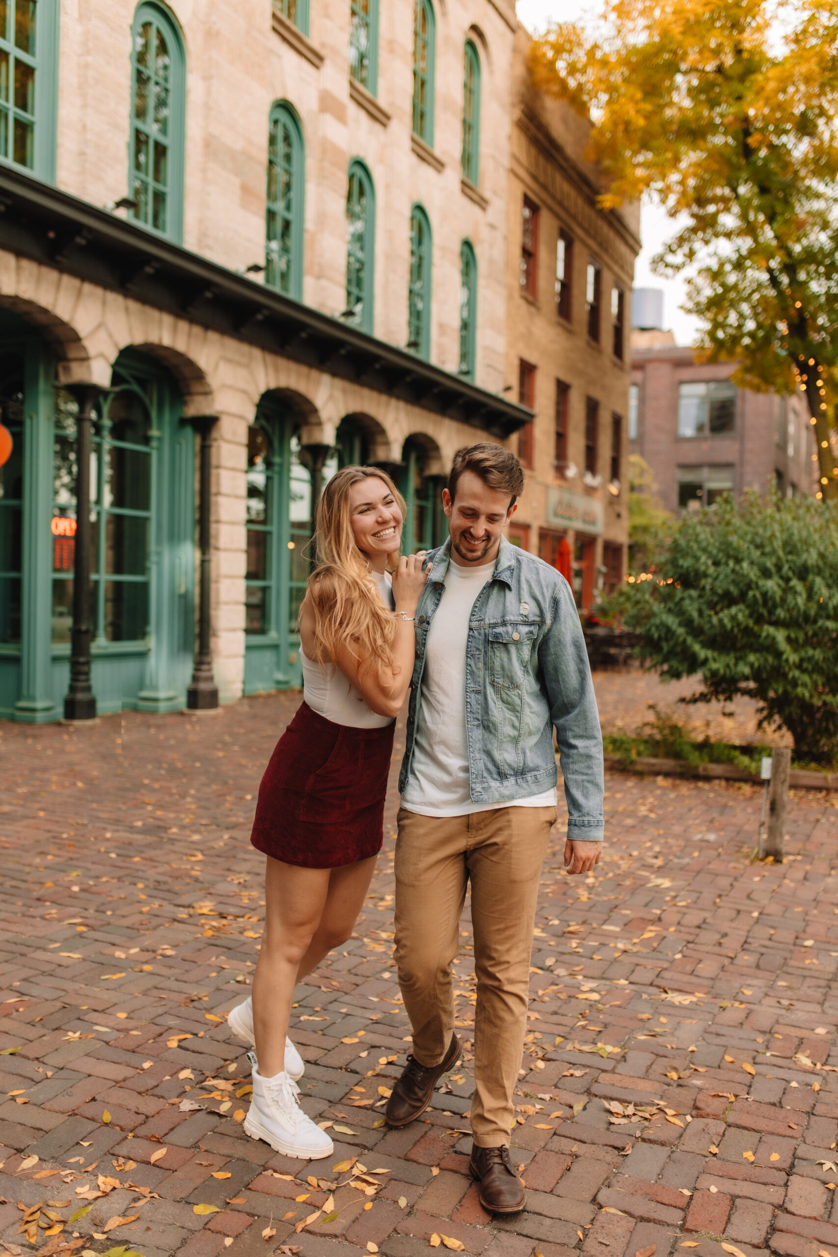 A young couple smiles and walks together on a brick pathway in a charming urban area with historic buildings and autumn foliage.