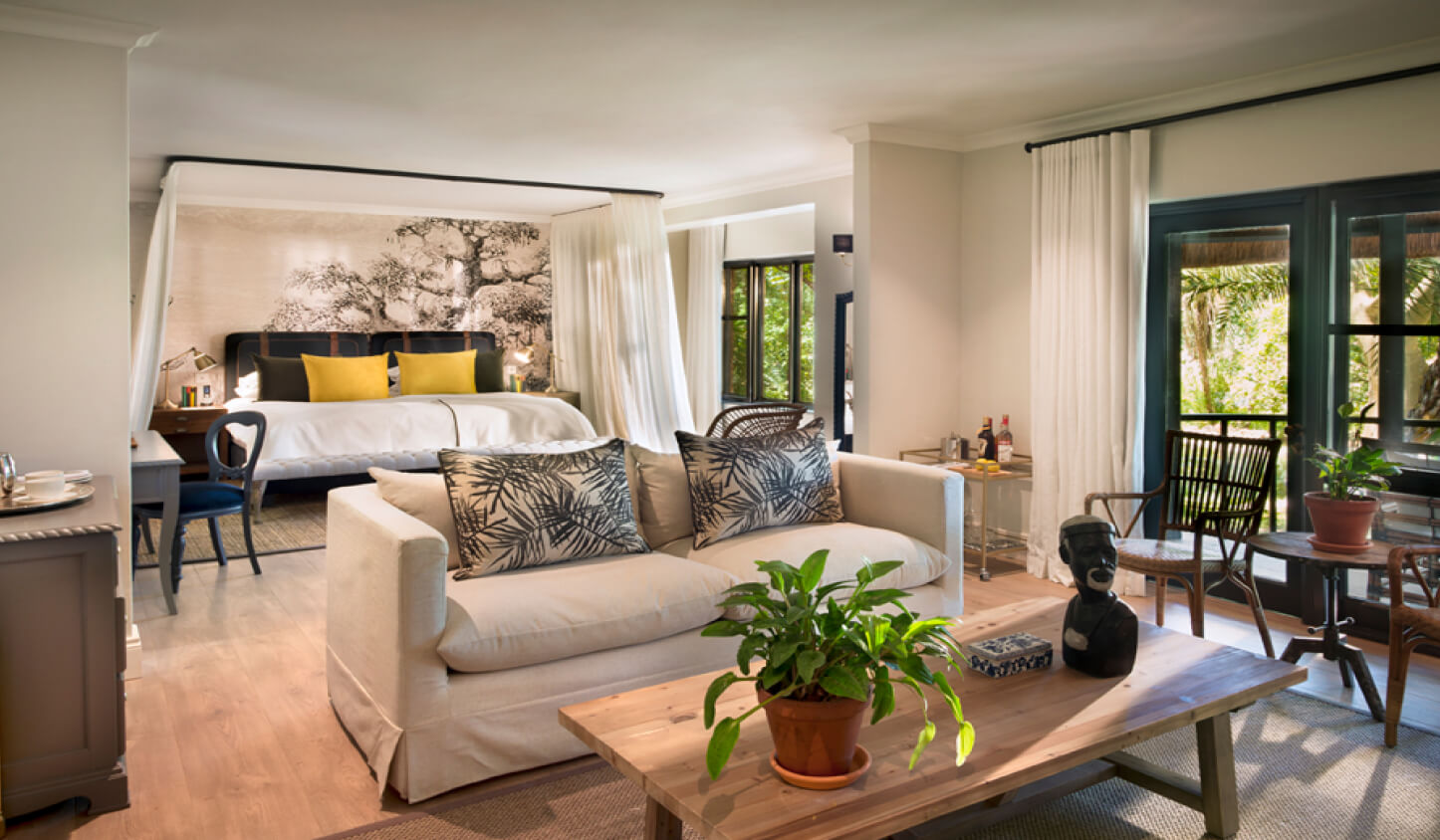 A spacious bedroom with a canopy bed, a seating area with a white couch and coffee table, indoor plants, and large windows with curtains, offering a view outside at lion sands perfect for a safari