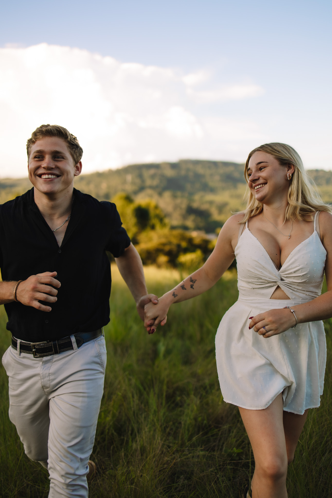 Newly engaged couple running holding hands smiling in a field of grass