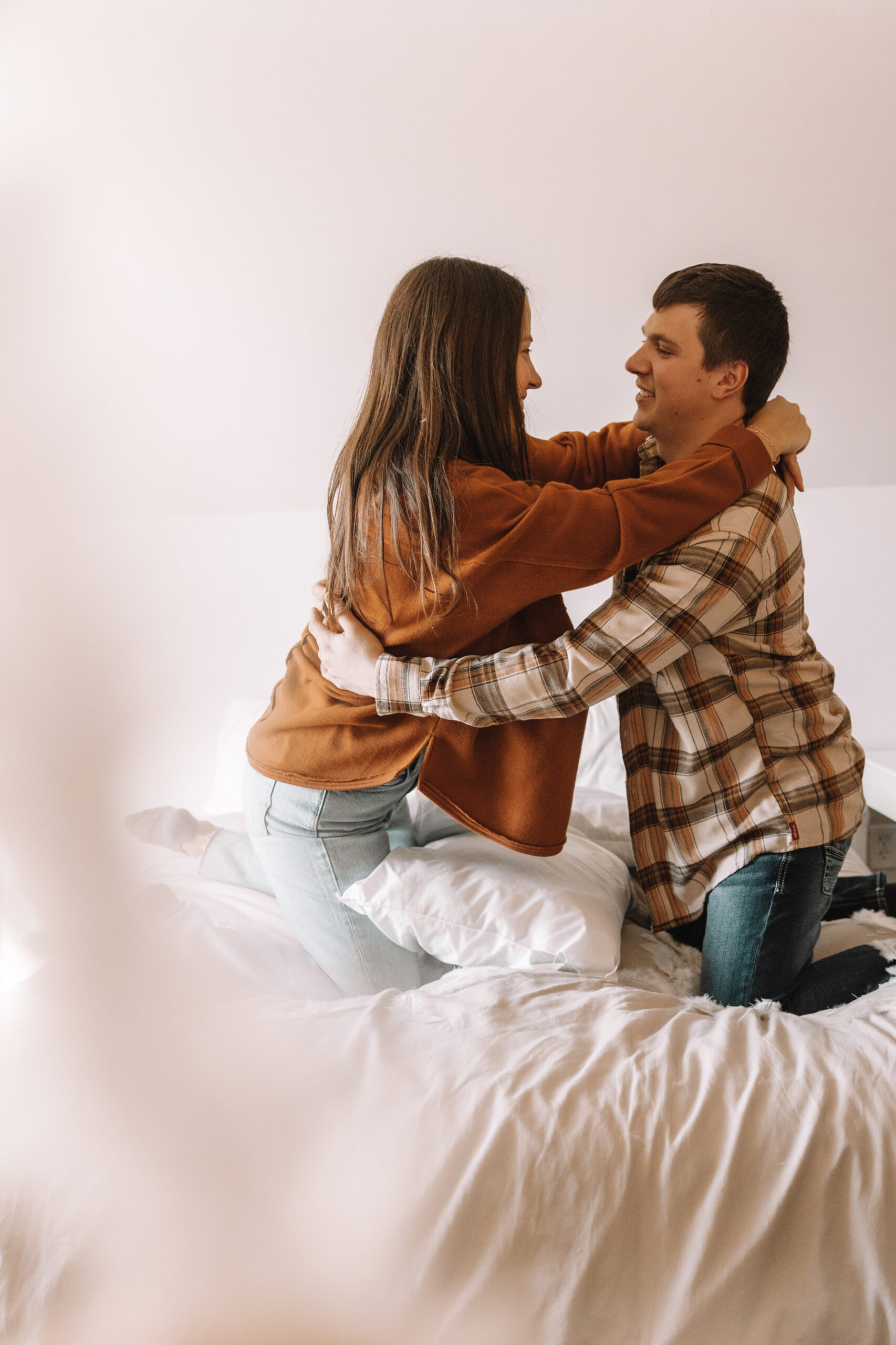 Newly engaged couple pillow fighting on a bed in long sleeve button ups and jeans for their unique engagement photo locations in Minnesota