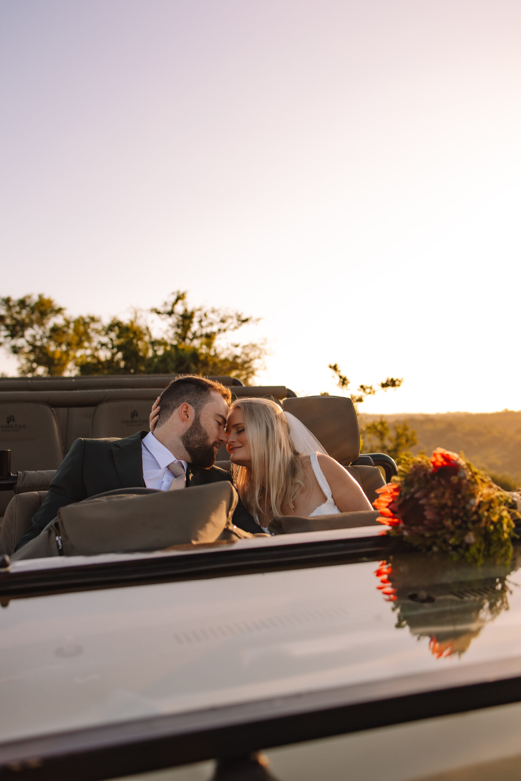 A bride and groom leaning their heads together while sitting in a safari vehicle during sunrise