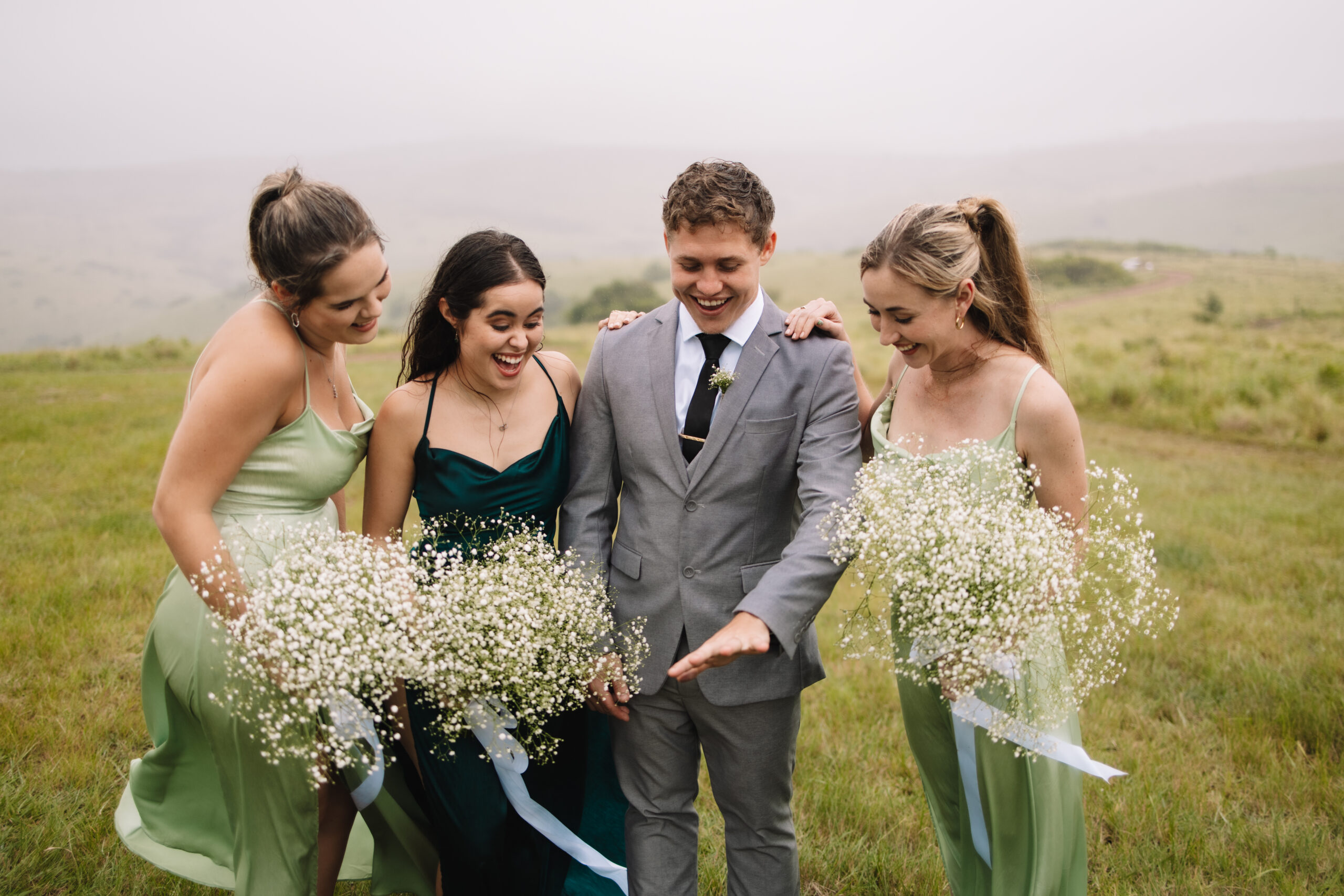 Groom showing off his ring to the bridesmaids as they stand and gasp while holding their bouquets consisting of babies breath flowers