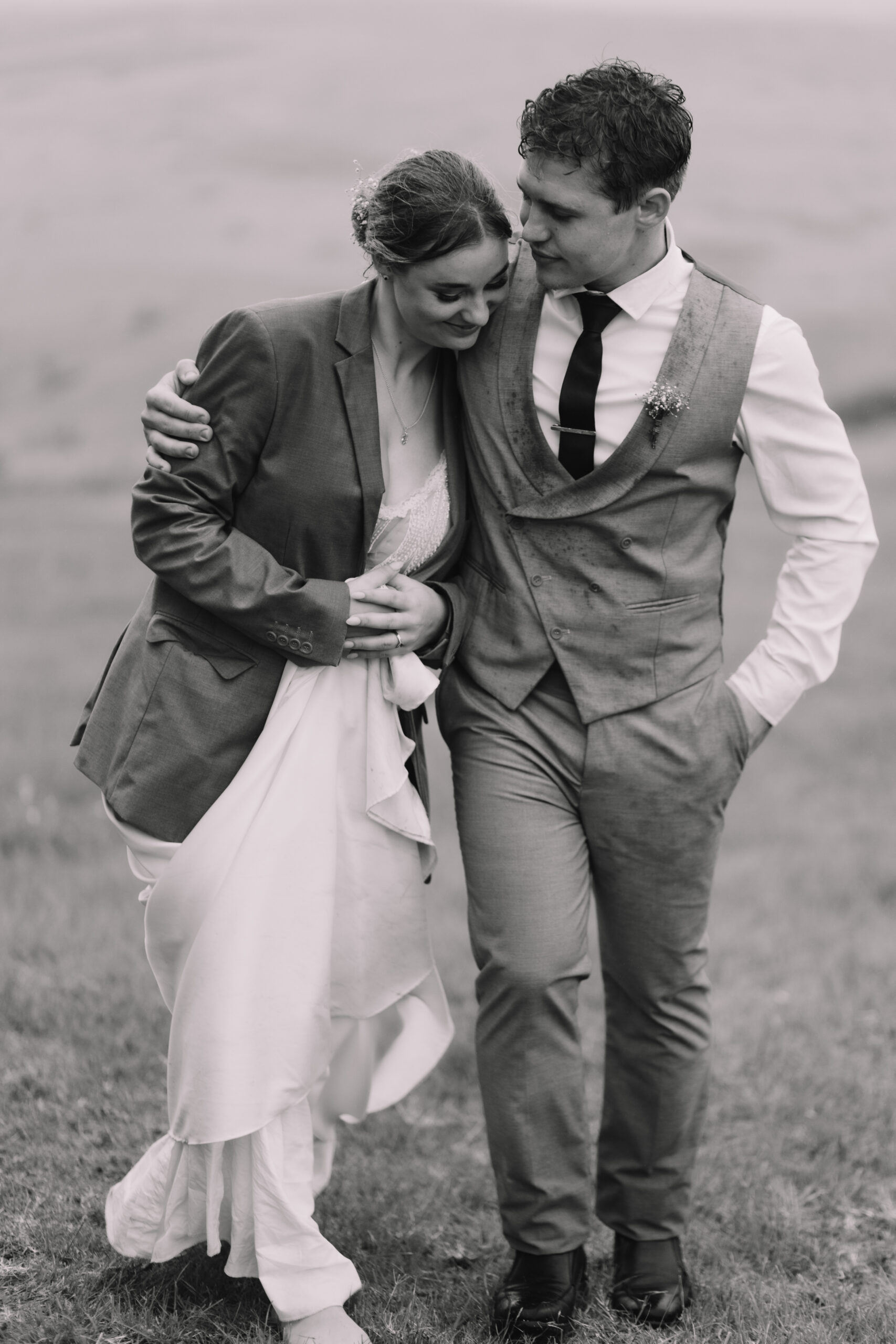 Black and white photo of bride wearing grooms suite jacket while they walk in a field of grass