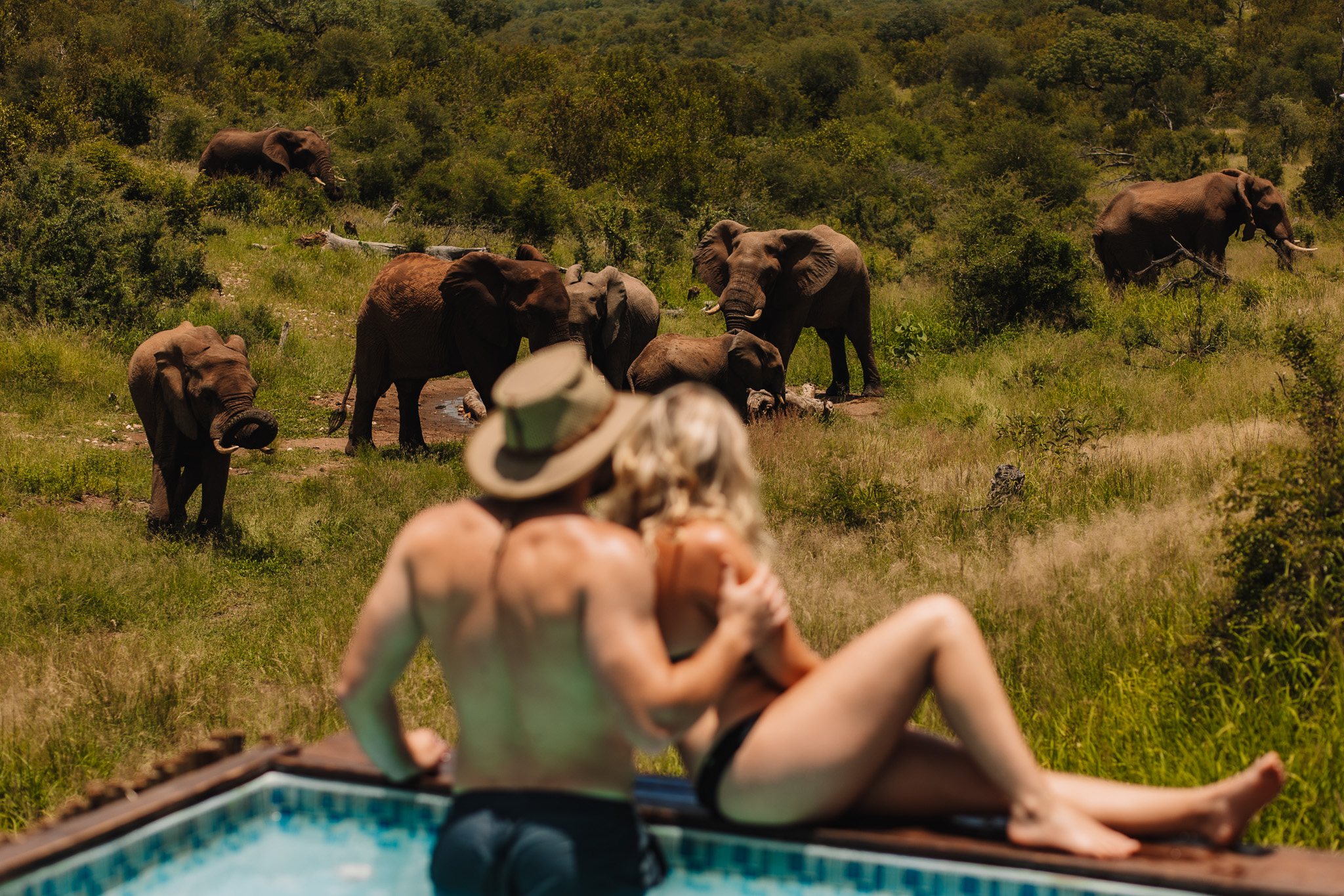 A model couple sitting by the pool at a safari lodge overlooking a watering hole with elephants, during a photography content retreat