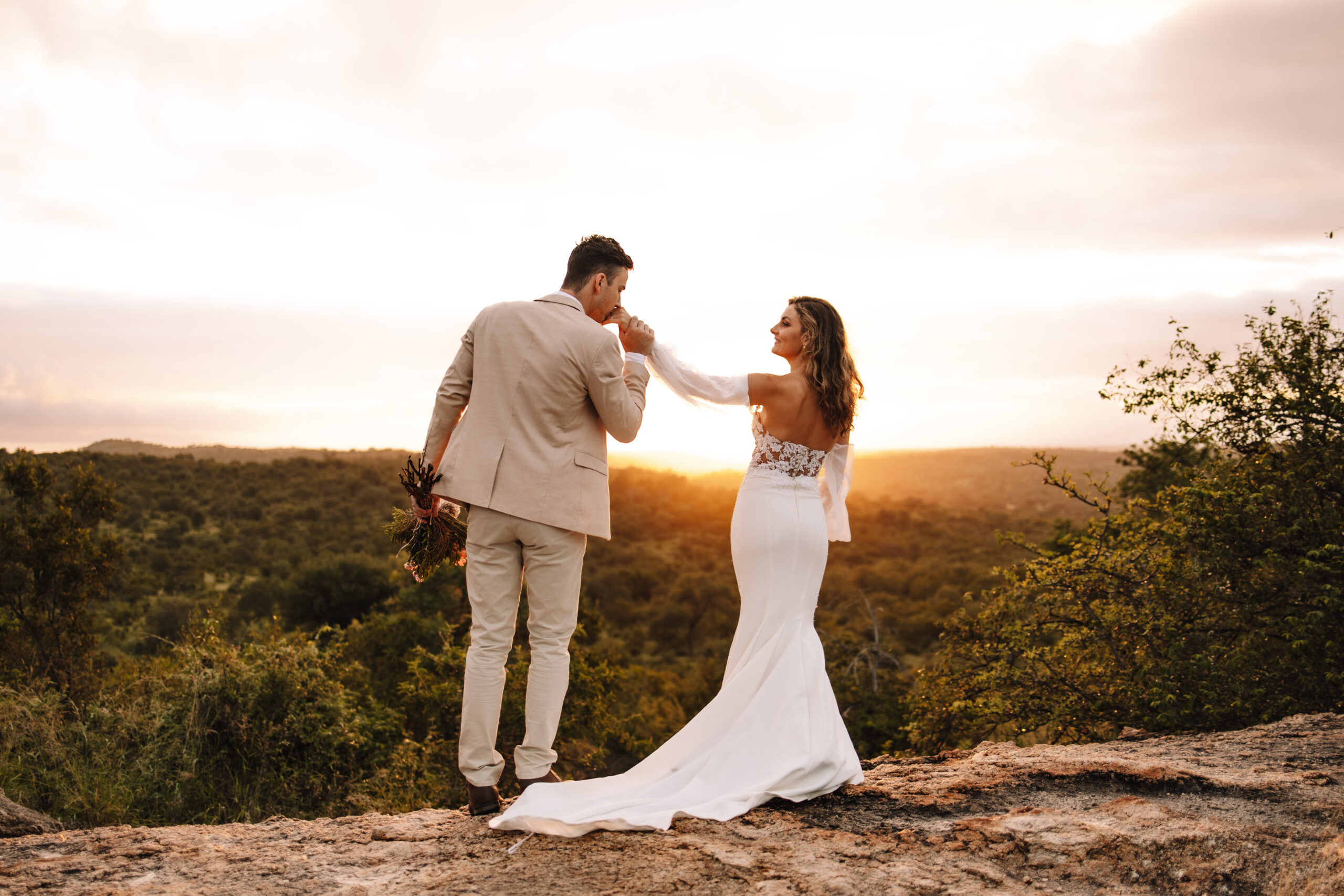 Destination wedding couple standing on the edge of a cliff while the groom takes his bride's hand and kisses it. The sun is setting behind them and the bride is wearing a beautiful open back, fitted wedding dress