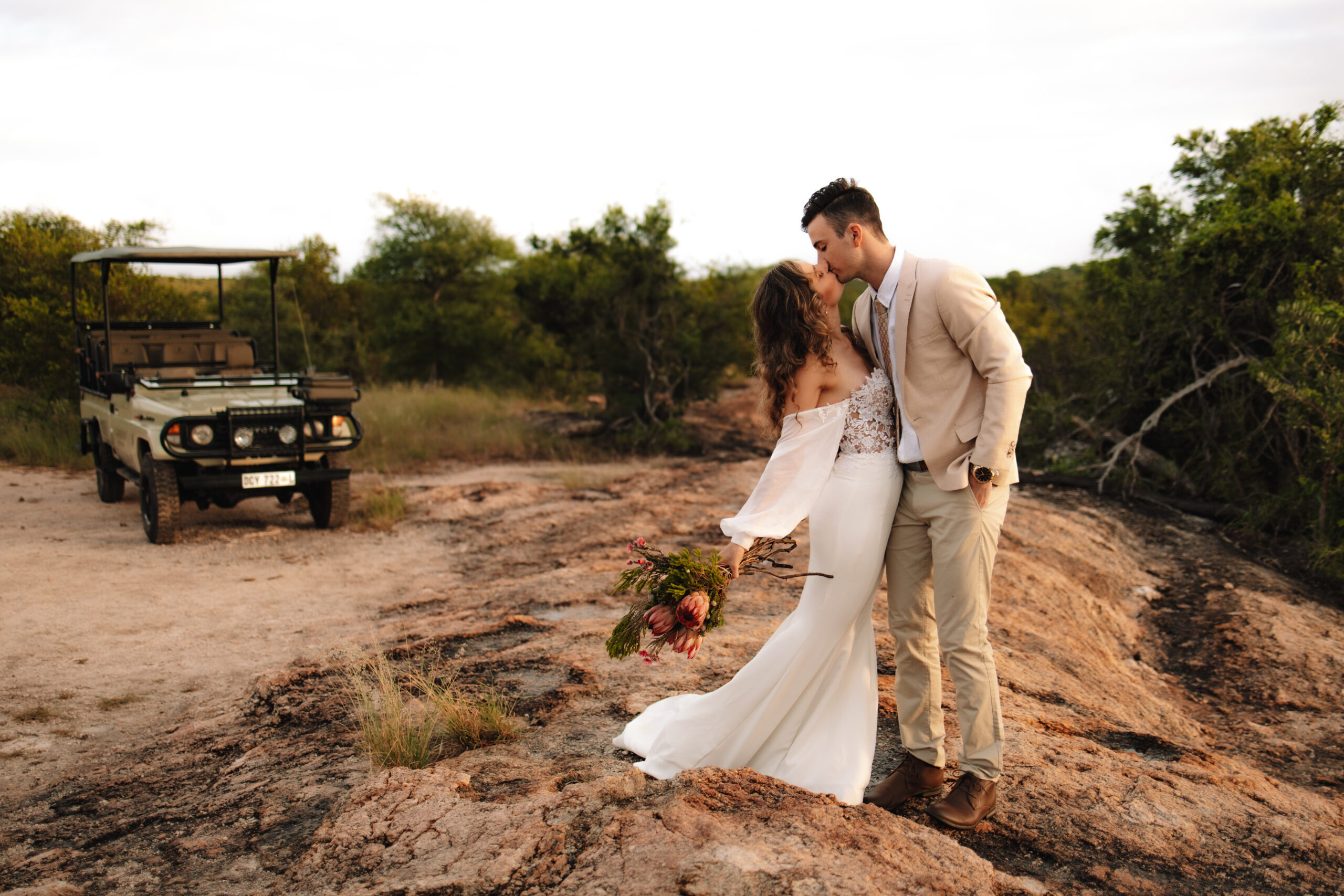 Bride and groom kissing while standing on a cliff with a safari vehicle behind them during a photography content retreat in Africa