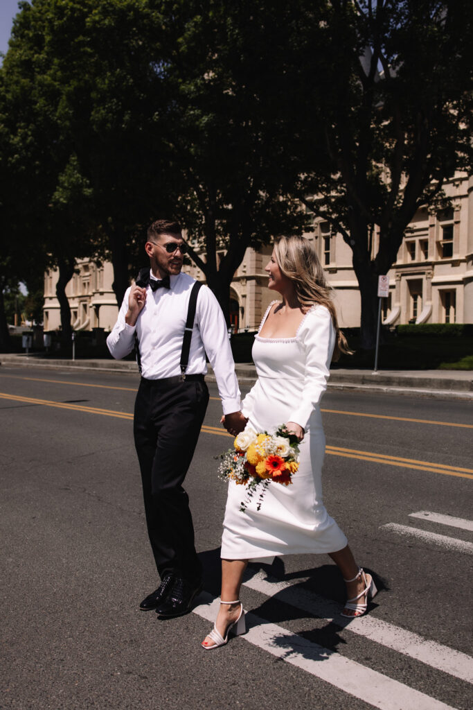 Bride and groom walking across the street holding hands. Groom is holding his suit jacket over his shoulder with his sunglasses on