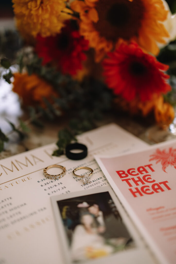 Detail flat lay shot of bride and groom's rings, bright summer flowers, a cocktail menu and a polaroid of a courthouse elopement