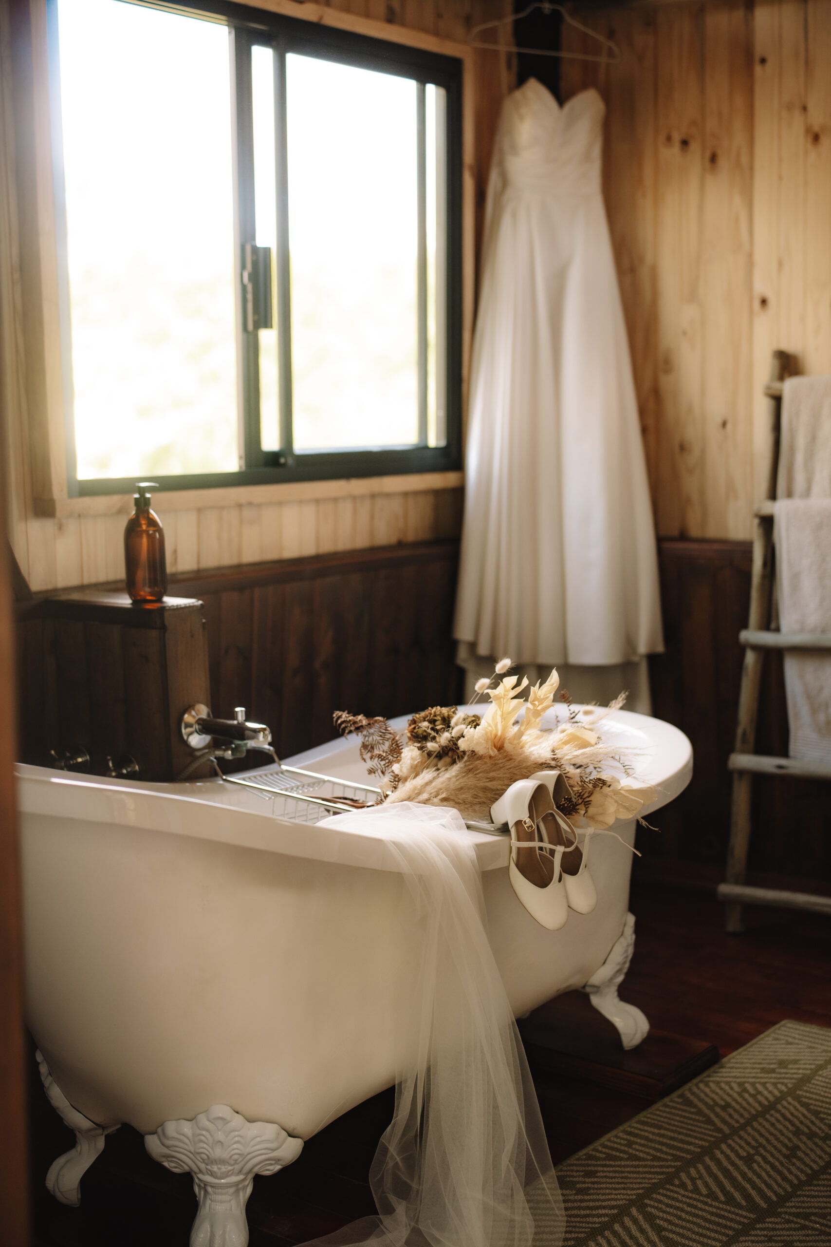 Beautiful safari wedding detail photo of veil, shoes and boho bouquet leaning over the bath tub with the wedding dress hanging in the background