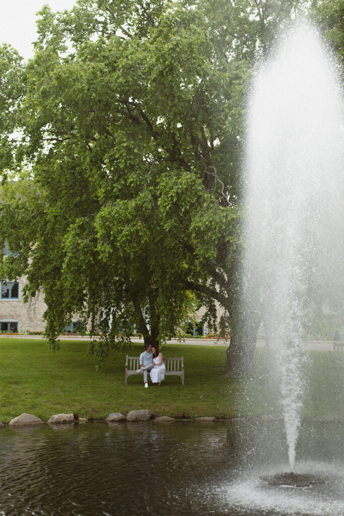 Engagement couple sitting on a bench next to a pond with a fountain for their engagement photos at the Minnesota Landscape Arboretum