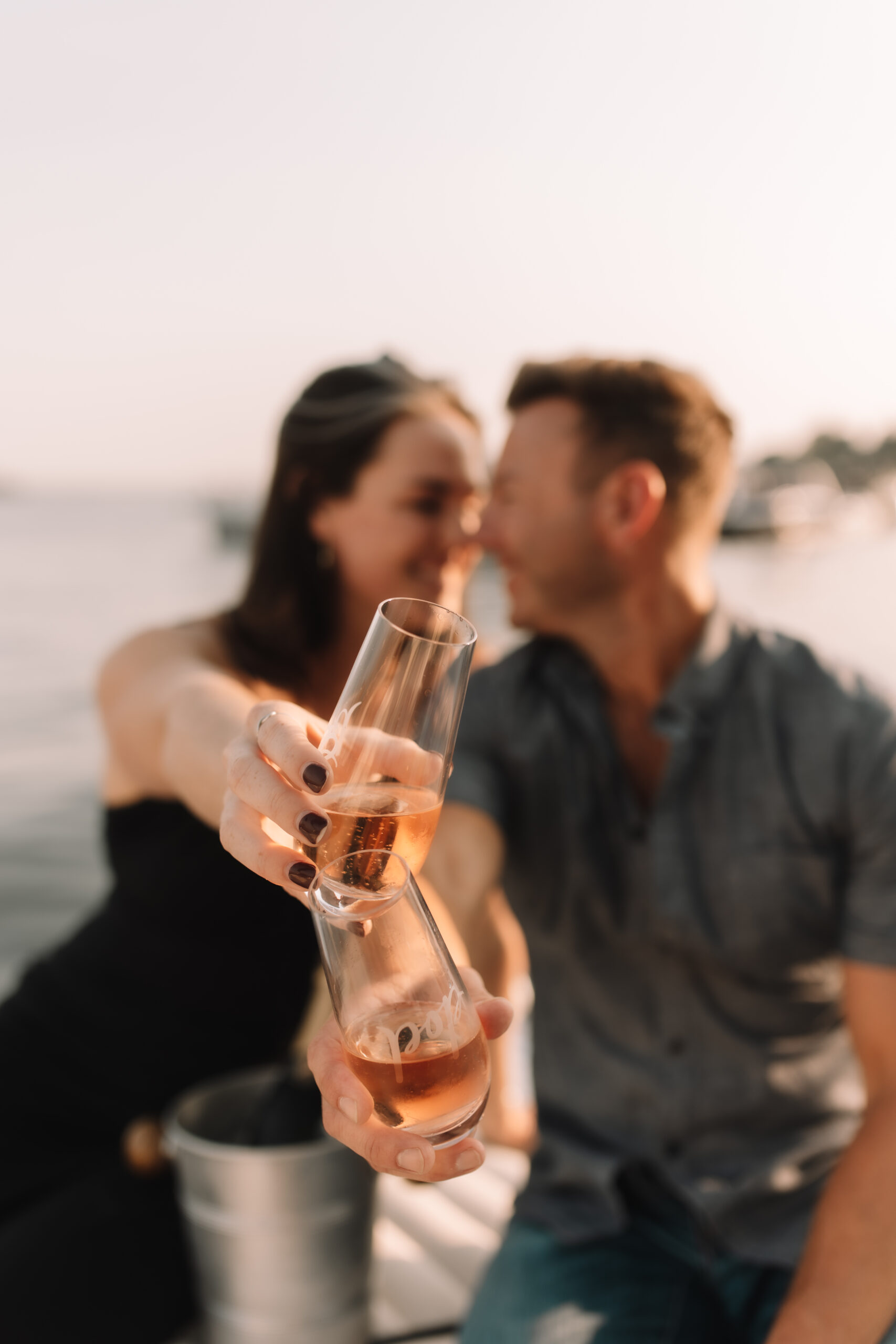 Guy and girl clinking their champagne glasses together during their engagement session at Wayzata Beach- one of the best engagement photo locations in Minneapolis' west suburbs