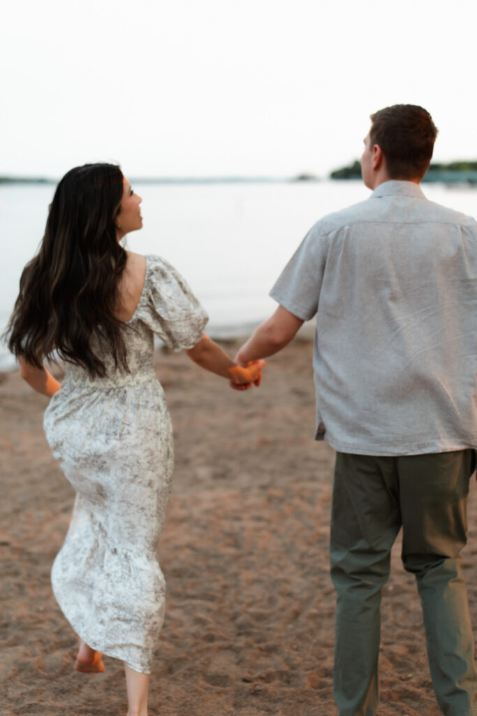 engagement couple holding hands and running on the beach towards Lake Minnetonka for their Wayzata Engagement Photos