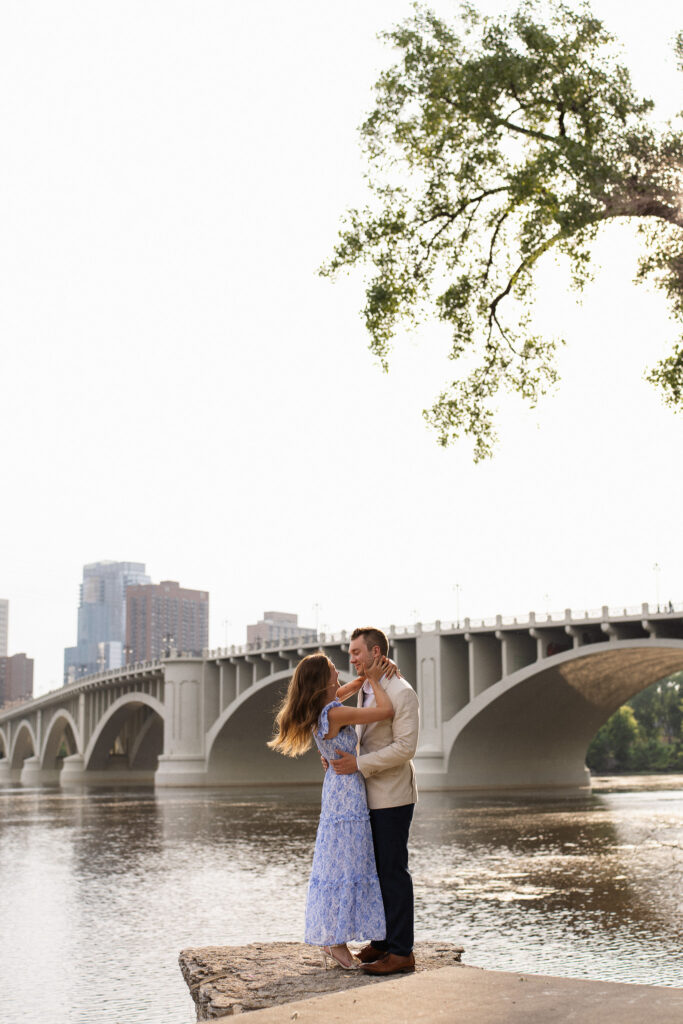 Engagement couple standing in front of the Minneapolis Skyline iconic view across St Anthony Main