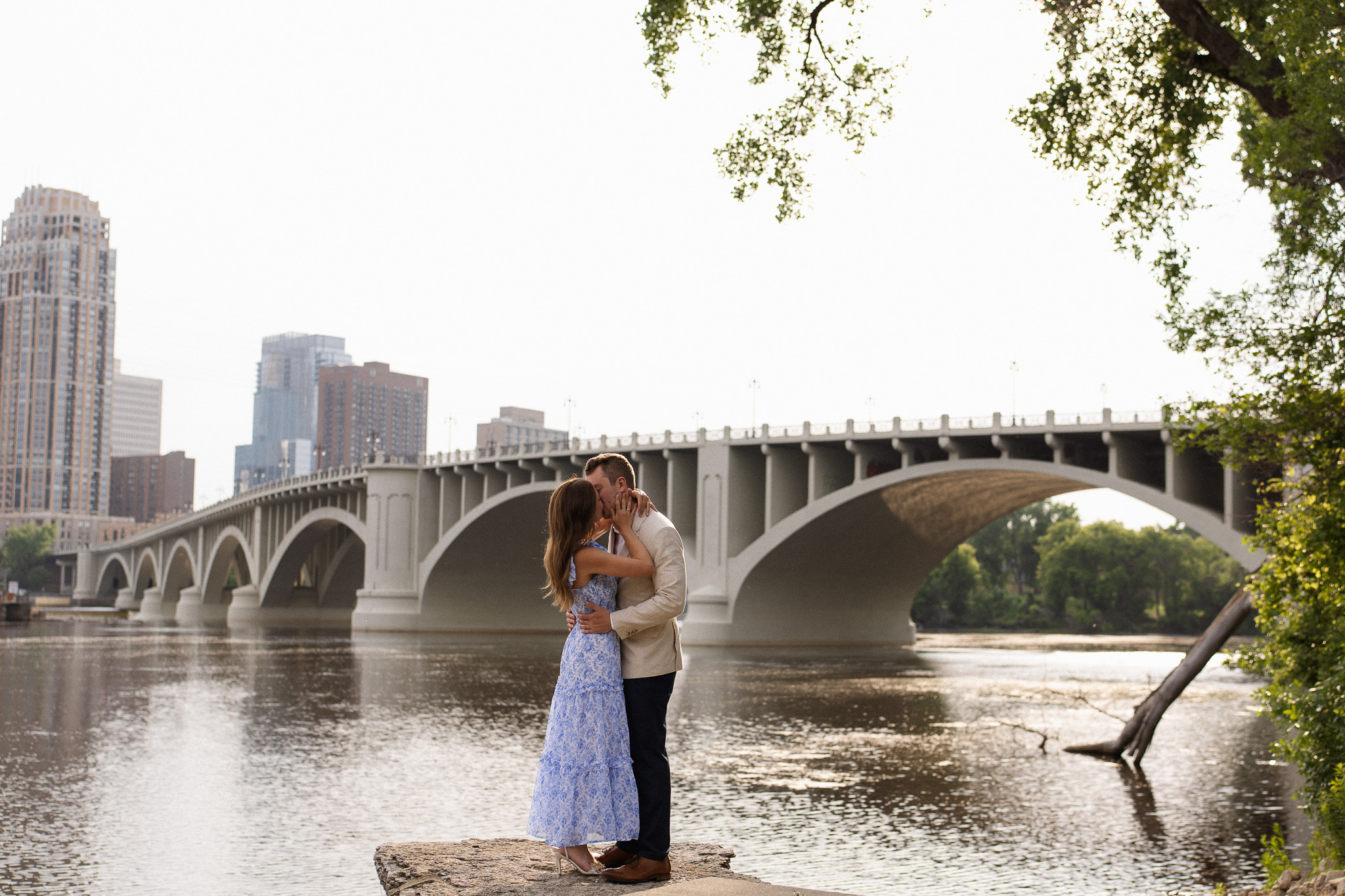 Minneapolis Engagement couple kissing in front of the bridge crossing the Mississippi river with the Minneapolis Skyline in the background