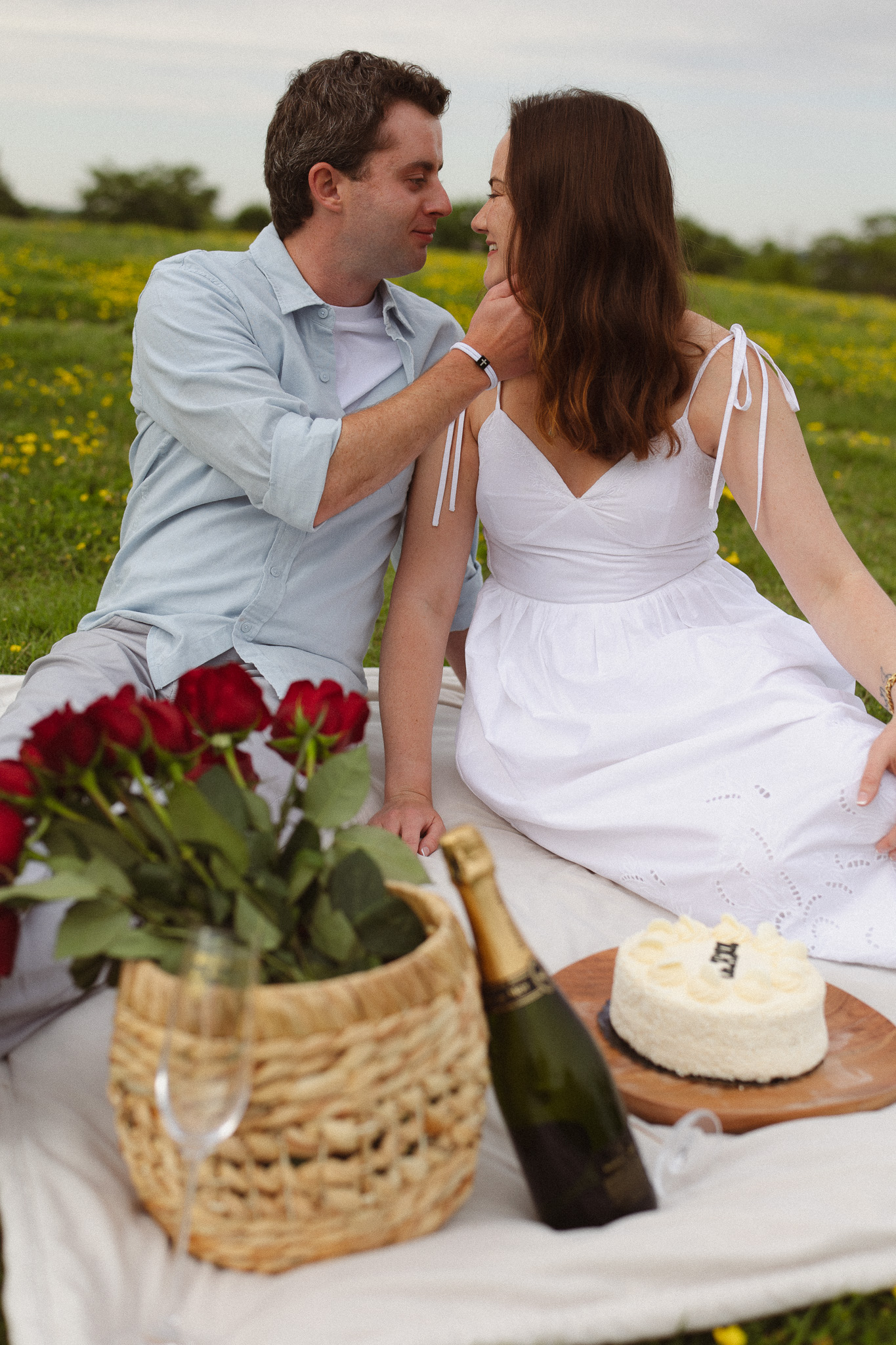 Couple sitting on a blanket having a pic-nic with props like roses in a basket, cake and champagne to celebrate their engagement