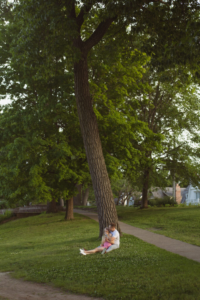 Wide shot of a couple sitting under a tree holding each other during their engagement photos in Excelsior